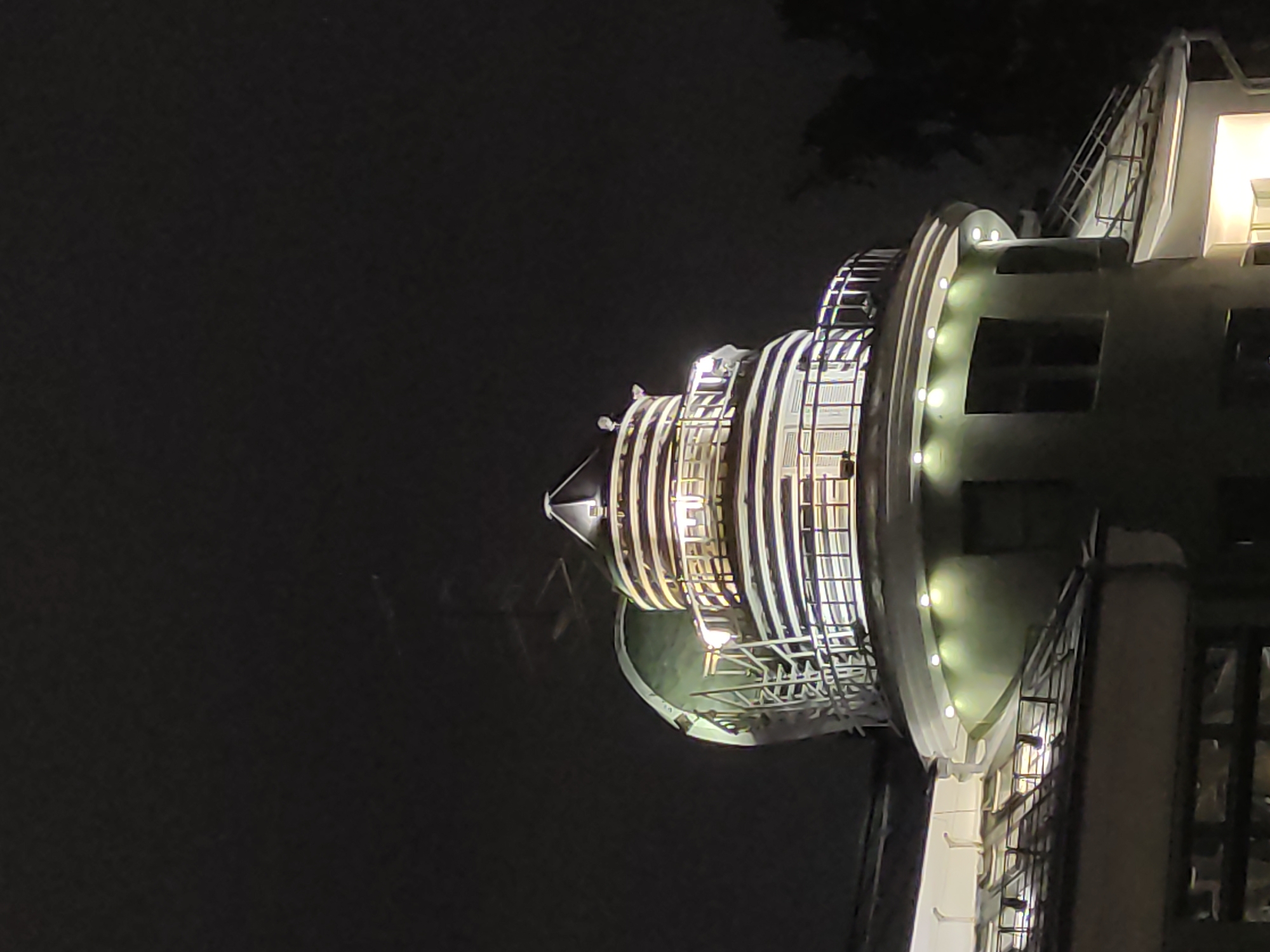 The image depicts the illuminated control tower of the Moscow State University at night. The tower features a multi-tiered, circular design with visible metal scaffolding and architectural lighting. Dark sky and tree outlines are visible in the background.