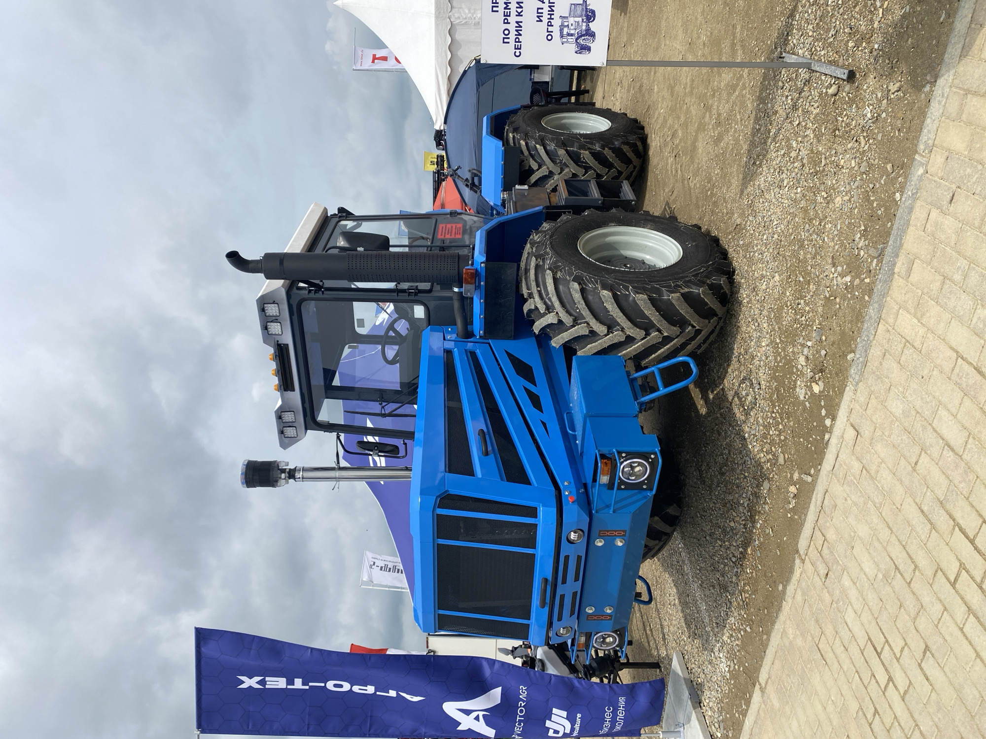 The image shows a blue tractor on display in Voronezhskaya, Russia. The tractor has large tires and a modern design. It is parked on a gravel surface next to a brick walkway. A banner with the text "APPO-TEX" is partially visible. The sky is cloudy. A sign with Russian text is visible in the background.
