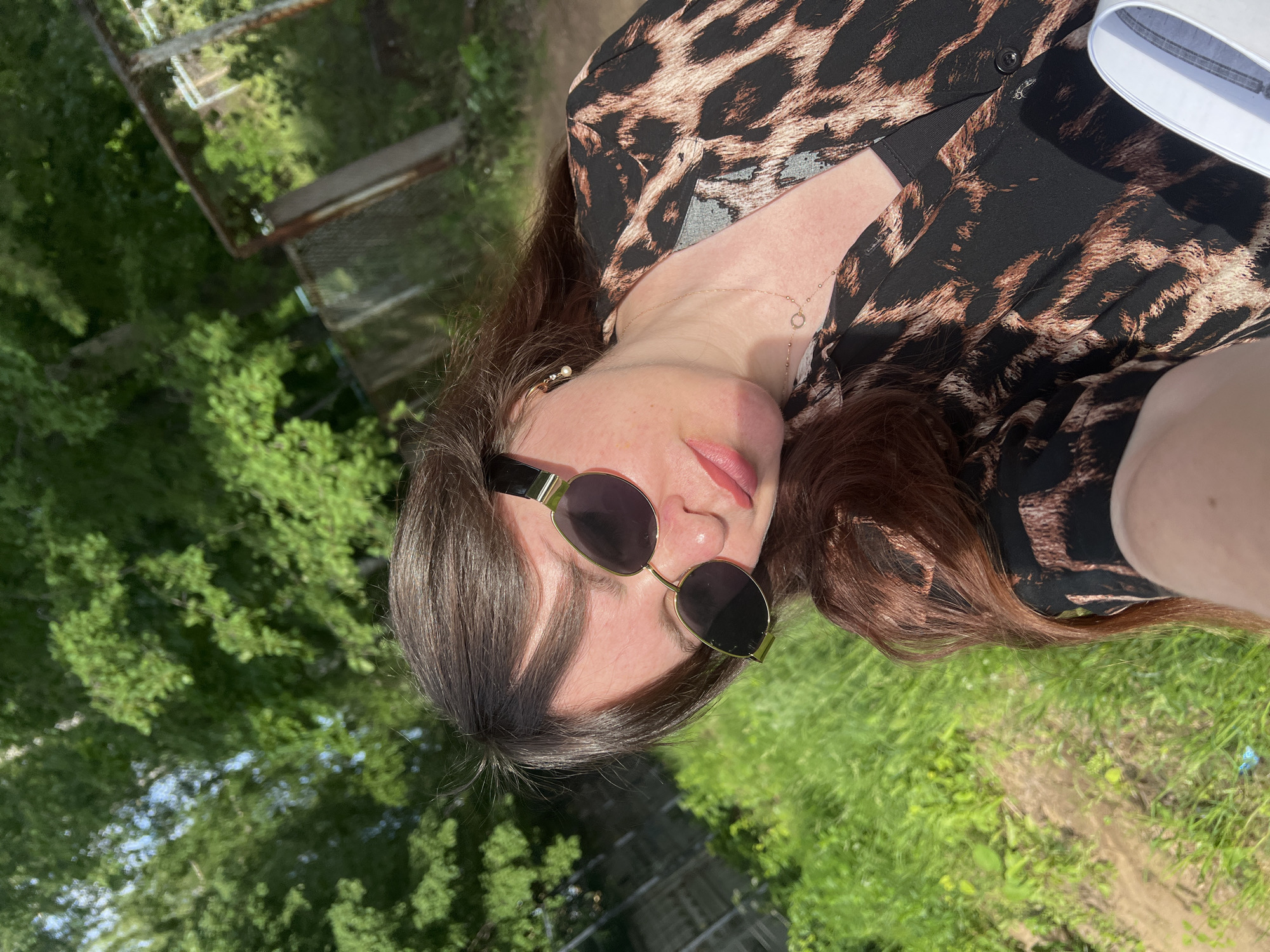 A woman with long, dark brown hair and fair skin is taking a selfie outdoors. She is wearing sunglasses with gold rims and a black and brown leopard-print button-down shirt over a black tank top. She has a delicate gold necklace with a small circular pendant. The background is blurred, showing lush green foliage, trees, and a rusty metal fence. The image appears to have been taken during the daytime, with sunlight illuminating the scene. The environment suggests a rural or semi-rural setting, possibly a backyard or a park, consistent with the outskirts of Noviy, Russia.