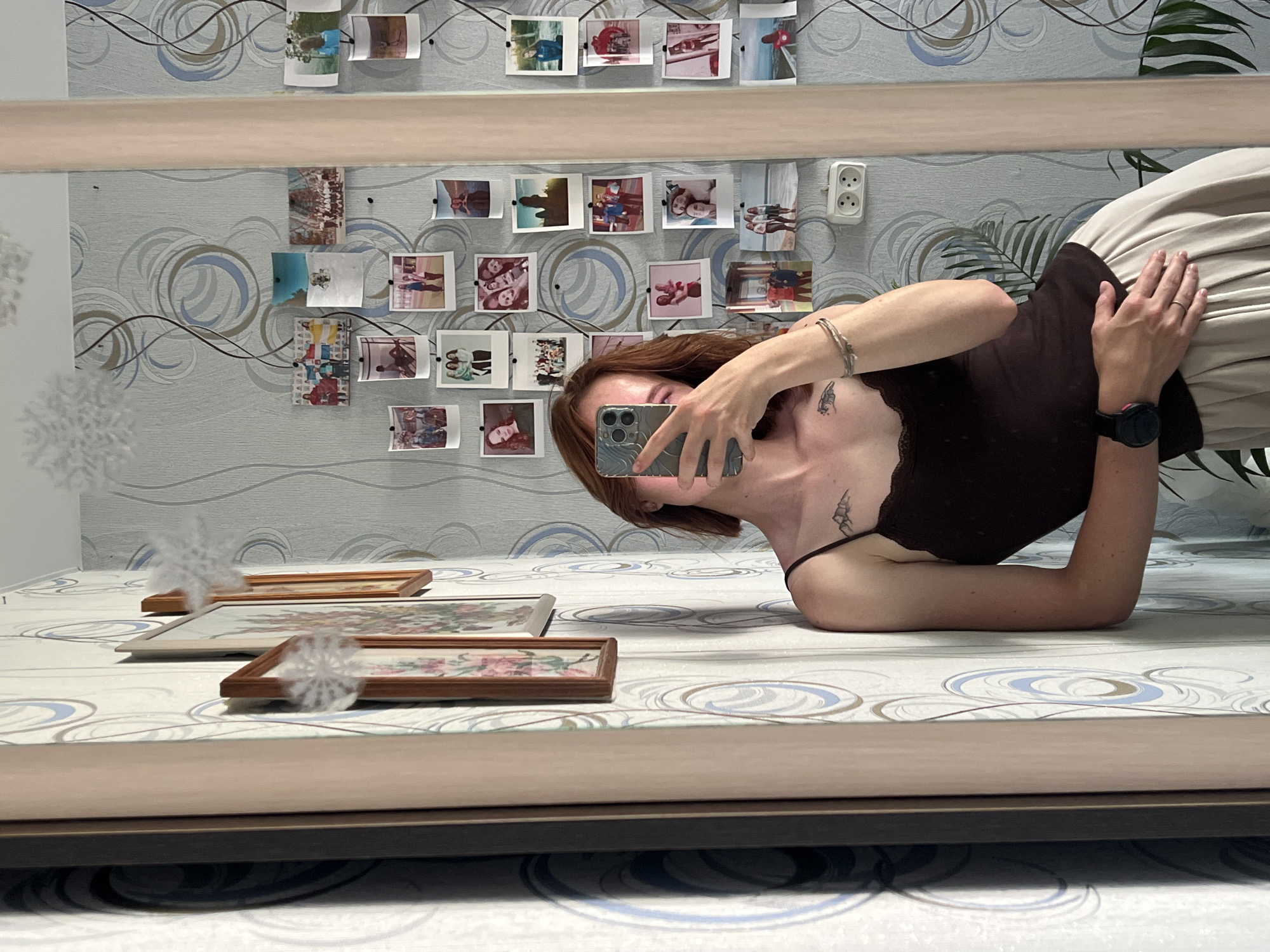 In a dimly lit room, a young woman with auburn hair is captured in a mirror's reflection, lying on a bed. She is wearing a dark brown camisole and a light-colored skirt. Her right arm is extended, holding a smartphone to take the photo, and her left arm rests behind her head. A tattoo is visible on her left upper chest and another on her right arm. She wears a bracelet on her left wrist and a smartwatch on her right.
The background consists of a wall adorned with a collection of small photographs pinned up, some showing people and others landscapes. A double electrical outlet is visible on the wall. Two framed pictures, one of flowers and the other with a floral pattern, are placed on the bed. The room appears to be a bedroom, suggested by the bed and the overall casual atmosphere. There is no indication of the time of day or weather conditions. The mood seems relaxed and personal, perhaps a moment captured for personal memory or social media. The location is specified as Chaikovsky, Russia.