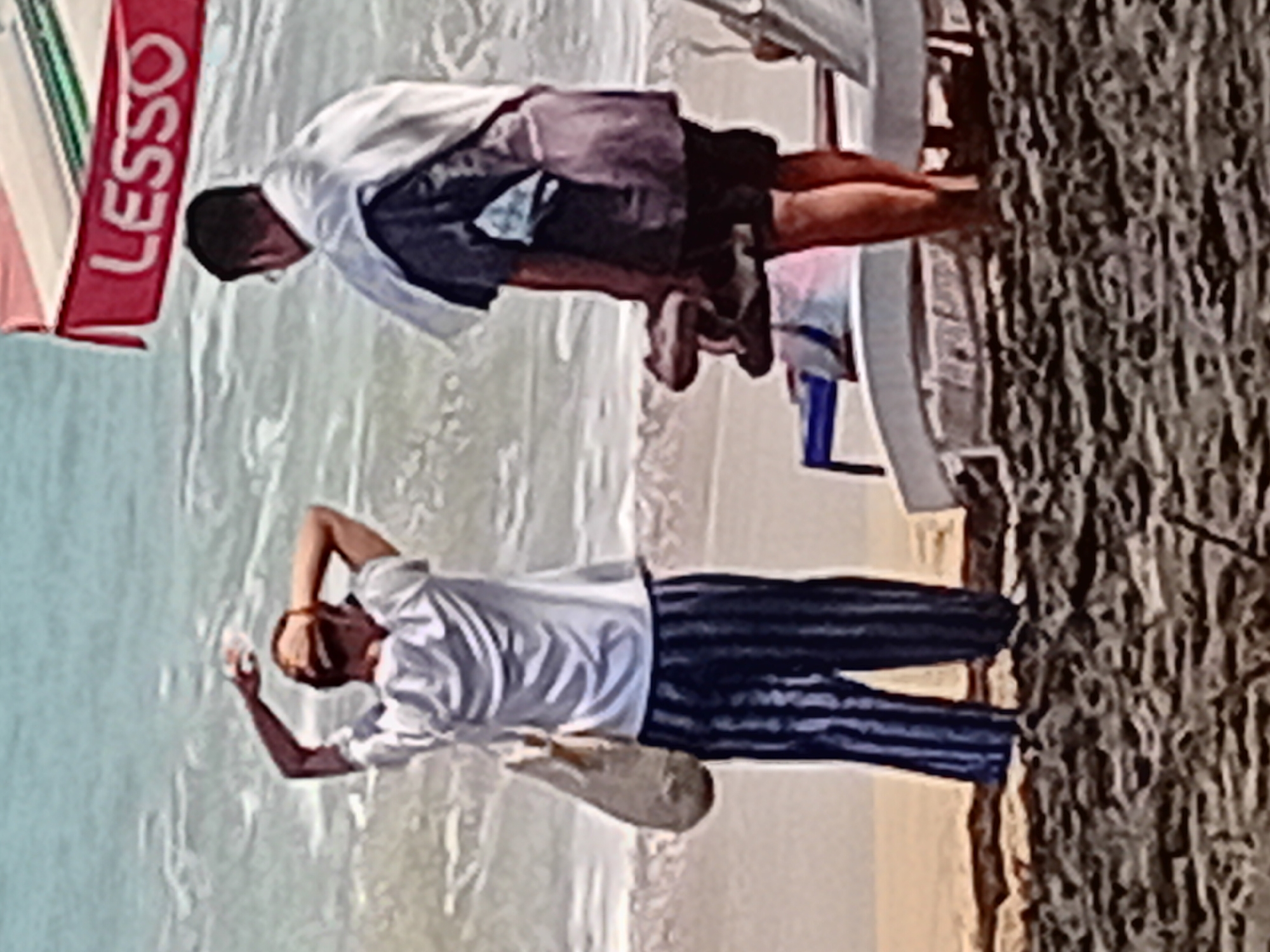 Two individuals are standing on a beach in Kuta, Indonesia. The person on the left, facing away from the viewer, is wearing a white t-shirt, striped pants, and a cap. They are holding a bag and have their hands on their head. The person on the right, also facing away, is wearing dark shorts and a shirt, with a white garment draped over their shoulders. They are holding a pair of sandals. In the background, the ocean waves are visible, and a red banner with "LESSO" written in white lettering is attached to an umbrella. The ground is covered in sand and pebbles.