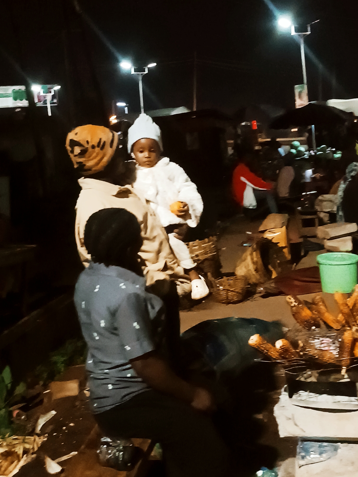 At night in Bukuru, Nigeria, a woman with an orange head covering holds a child wearing a light blue hat and white clothing. The child holds an orange object. A person with short dark hair sits facing away, wearing a patterned blue shirt. Roasted corn is visible nearby. Streetlights illuminate the scene. Woven baskets and a green bucket are on the ground. Other people are sitting in the background.