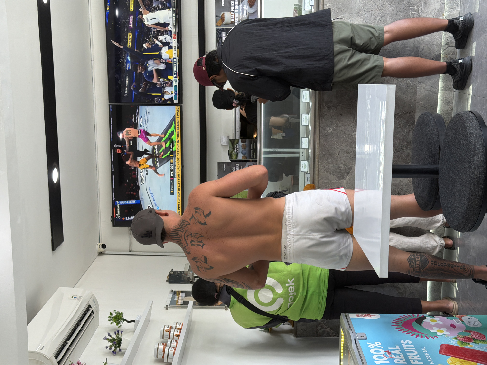 An indoor scene depicts several people near a counter with food items and televisions displaying sports. In the foreground, a man with extensive back and leg tattoos is wearing a baseball cap and white shorts. To his left, a person in a bright green vest with a "Gojek" logo is visible. To the man's right, another person wearing a dark jacket and a maroon cap is looking towards the counter. The environment appears to be a shop or eatery, possibly a cafe or convenience store, with shelves displaying products and a refrigerated display case. The televisions show a mixed martial arts fight and a basketball game. The overall impression is a casual setting where patrons are observing sports while potentially purchasing items.
