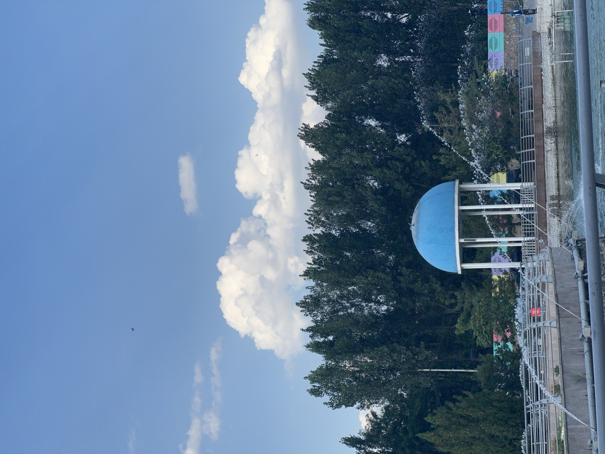 This image depicts a park scene, likely in Shymkent, Kazakhstan, characterized by a bright blue sky with scattered white clouds and two small birds in flight. In the foreground, a body of water with active fountains is visible, with metal railings and pipes surrounding it. A prominent blue domed structure, supported by white pillars, stands in the middle ground, partially obscured by greenery. Behind the structure, a dense line of dark green trees forms a backdrop. Further back, a row of colorful decorative panels, in shades of pink, yellow, green, and blue, are affixed to a fence. A person in a dark outfit and a blue cap stands near the fence, holding a hose, seemingly attending to the fountains. The overall impression is that of a well-maintained urban park on a clear day.