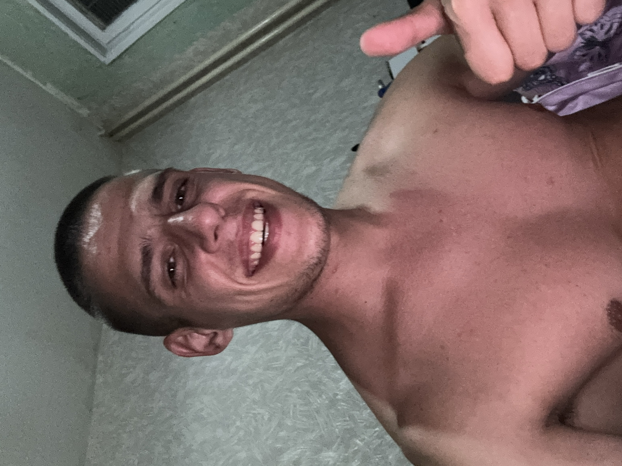A shirtless man with short, dark hair is smiling and giving a thumbs-up gesture. He appears to be indoors, with a patterned wall visible in the background. The location is undetermined as it could not be verified from the picture if it's in the city of Sofia, Bulgaria.