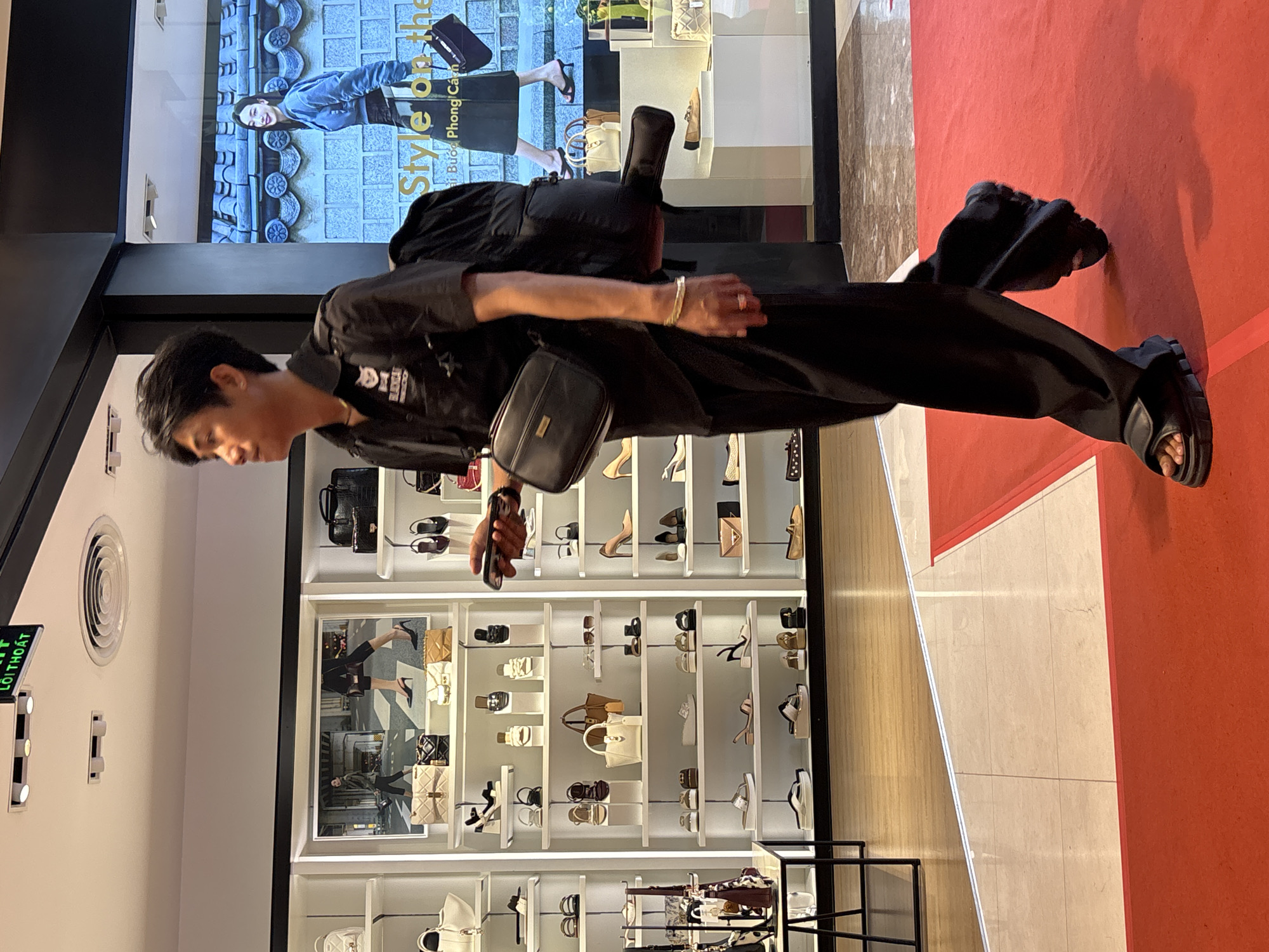 A young Asian man, wearing a black short-sleeved collared shirt with a patch on the left chest, and loose-fitting black pants, walks across a red carpeted area in a brightly lit retail environment. He has a black crossbody bag slung over his right shoulder and is carrying a black backpack on his back. His right hand is holding a smartphone. The background features shelves stocked with various women's shoes and handbags. Above the shelves, a large digital advertisement displays a woman in a blue jacket. The flooring transitions from wood to tiled surfaces. A sign at the top left corner reads "LÔI THOÁT" in green text. The scene is likely in a department store or a high-end boutique in Nha Trang, Vietnam, given the Vietnamese text on the sign and the overall appearance of the store.