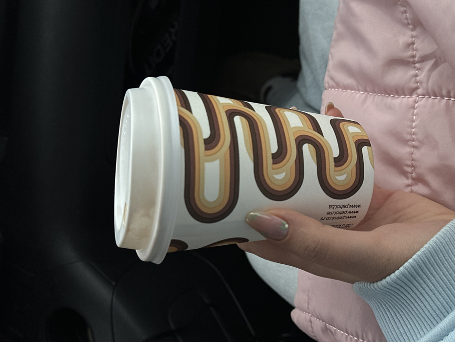 The image displays a close-up of a human hand holding a disposable coffee cup within what appears to be a vehicle's interior. The hand exhibits manicured fingernails with a light nude polish and subtle glitter on the tips. A light blue long-sleeved garment is partially visible on the arm.
The disposable cup is white, adorned with a repeating retro-style pattern of wavy, parallel stripes in shades of brown, beige, and light cream. Printed vertically on the cup are the web addresses "PETROL.CO.RS", "WWW.PETROL.ME", and "WWW.PETROL.BA". The cup is sealed with a white plastic lid, which shows slight brown residue on its interior surface, consistent with a coffee-like beverage.
Below the cup and hand, a light pink quilted fabric is visible. The background is dark and indistinct, characteristic of a car's interior. The branding on the cup identifies it as originating from a Petrol service station, which operates in Montenegro. However, the specific location within the city of Bartula, Montenegro, is not discernible from the visual information provided in the image.