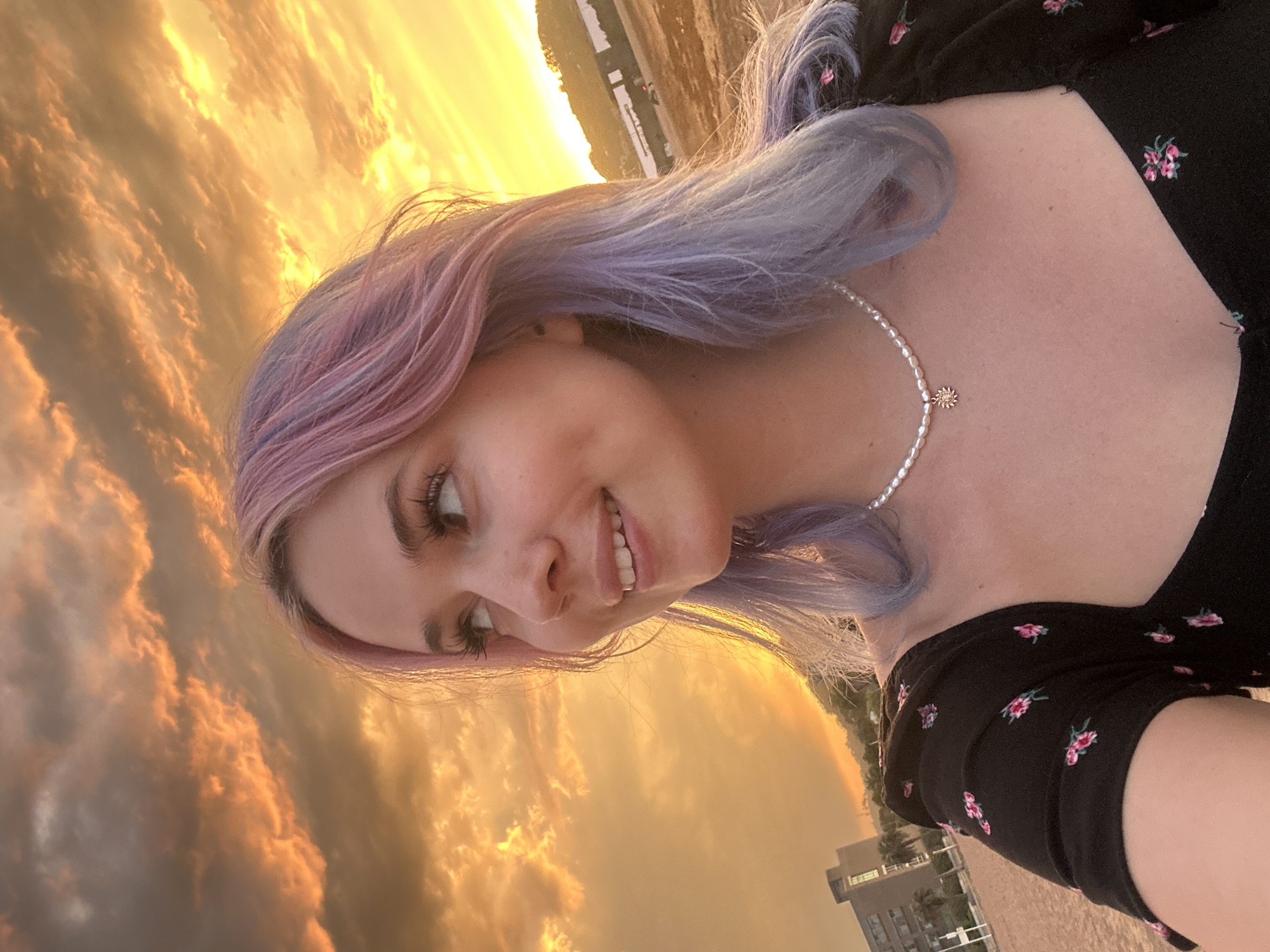 A young woman with multi-colored hair, styled in pink and lavender hues, smiles as she looks to her left. She wears a black dress adorned with a floral pattern and a pearl necklace with a sun-shaped pendant. The setting appears to be a beach in Gijón, Spain, with a building and a hillside visible in the background under a vibrant sunset sky. The sand on the beach is a light brown, and the water is not visible.