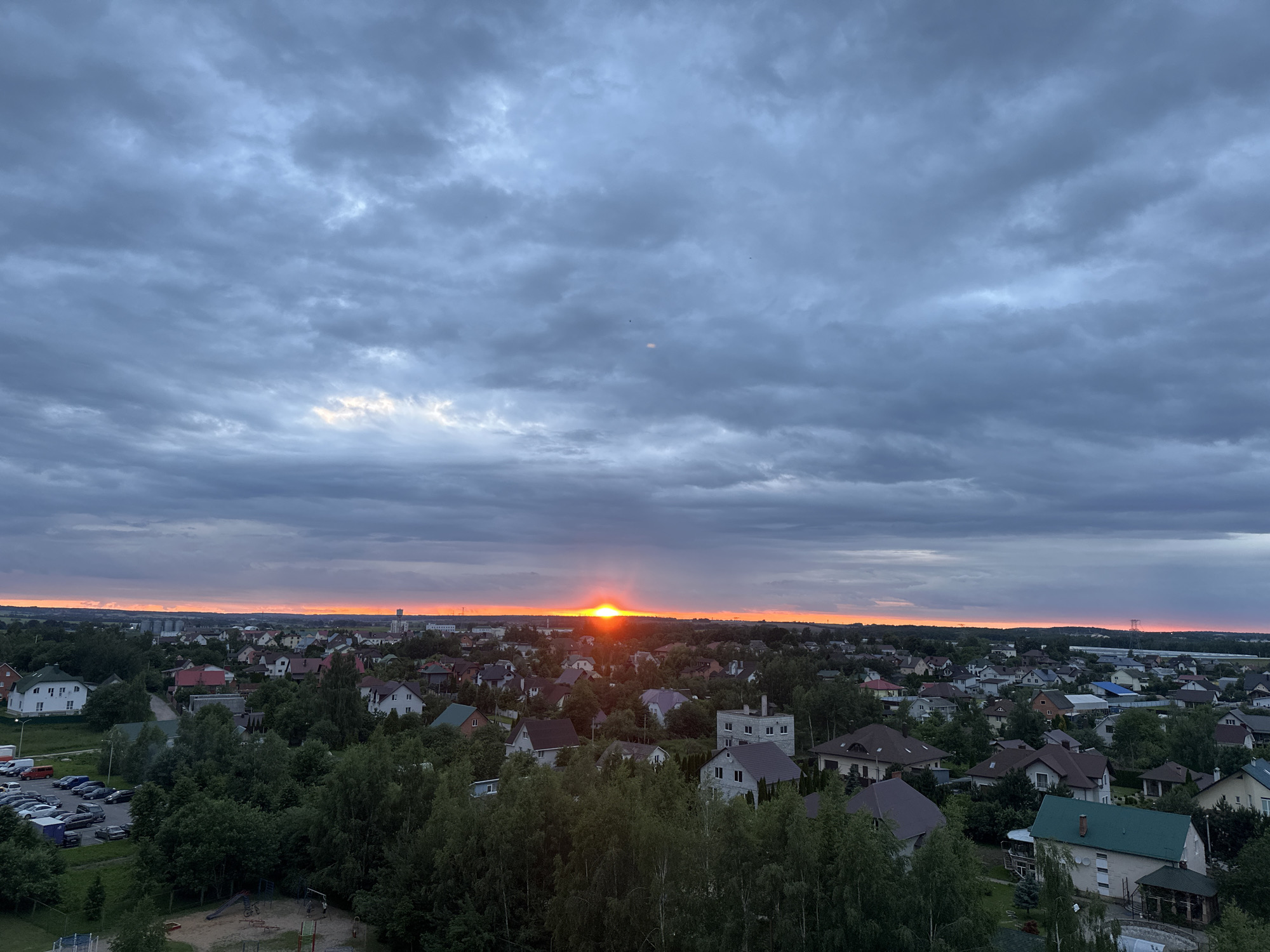 The image captures a panoramic view of a suburban area in Шчомысліцкі сельскі Савет, Belarus, during a sunset. The sky is filled with dramatic, grey clouds, with a bright orange glow emanating from the setting sun near the horizon. Below the sky, a dense collection of residential houses with varied roof colors and architectural styles covers the landscape, interspersed with numerous green trees. In the foreground, a playground area with swings and slides is visible, and to the left, a parking lot is filled with several cars. The scene depicts a peaceful, late evening in a residential district.