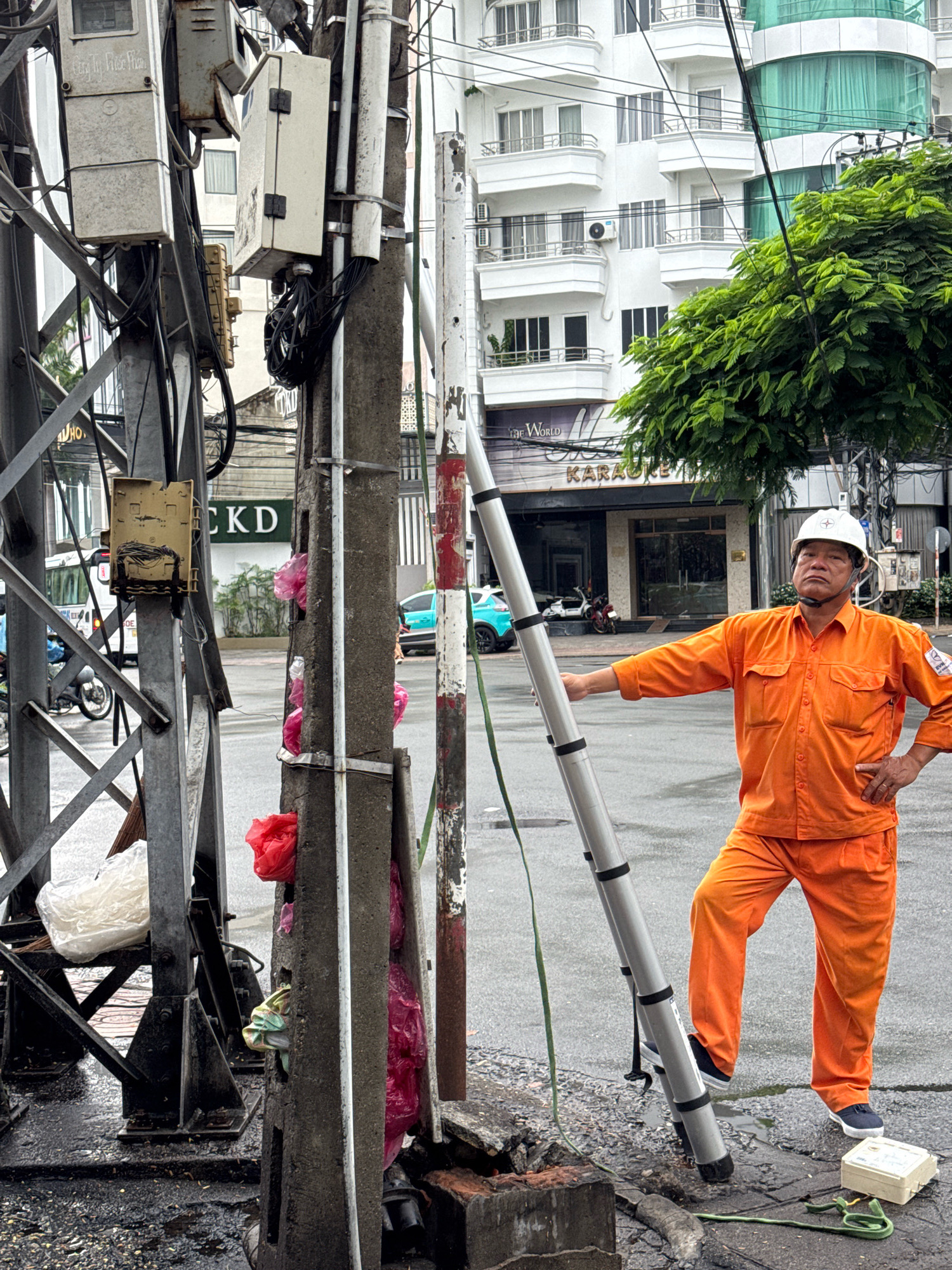 A male utility worker, wearing an orange uniform and a white hard hat, stands with his right hand on his hip and his left hand holding a long, segmented pole. He is looking towards the camera with a neutral expression. The worker is standing on a wet, paved street in front of a concrete utility pole and a large metal utility tower. The utility tower is covered in various electrical boxes and cables. Several brightly colored plastic bags are stuffed into crevices in the concrete utility pole. In the background, a modern white building with balconies and green tinted windows is visible, as well as a tree with dense green foliage. A turquoise car is parked on the street. The scene appears to be in an urban environment, likely Nha Trang, Vietnam, given the architecture and vegetation.