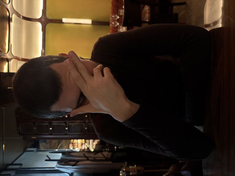 A man in a dark long-sleeved shirt sits at a table, his face in his hands. His head is angled towards the left with short dark hair. He appears distressed or contemplative. A lit bar area and other seating are visible in the background, suggesting he is in a restaurant or bar setting. A warm light casts a glow on the scene, and a dark wooden table is prominent in the foreground.