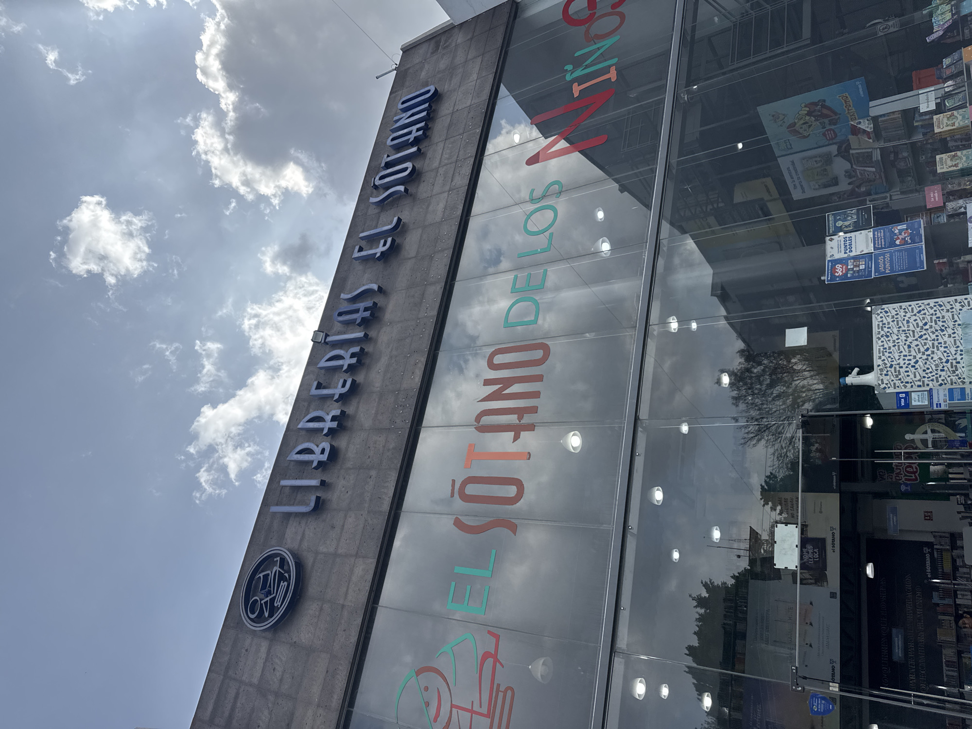 This photograph captures the exterior of Librerías El Sótano, a bookstore located in Puebla, Mexico. The image, taken from a low angle, highlights the store's signage. The main sign, "LIBRERIAS EL SOTANO," is displayed vertically in bold, blue letters. Next to it, "EL SÓTANO DE LOS NIÑOS" is vertically displayed on the window in colorful lettering, suggesting the store has a children's section. The glass windows reflect the sky and surrounding buildings, showcasing various advertisements and book displays. Inside, bookshelves laden with books are visible. Additional signs and advertisements are plastered on the windows and doors. The sky is partly cloudy, with sunlight filtering through.