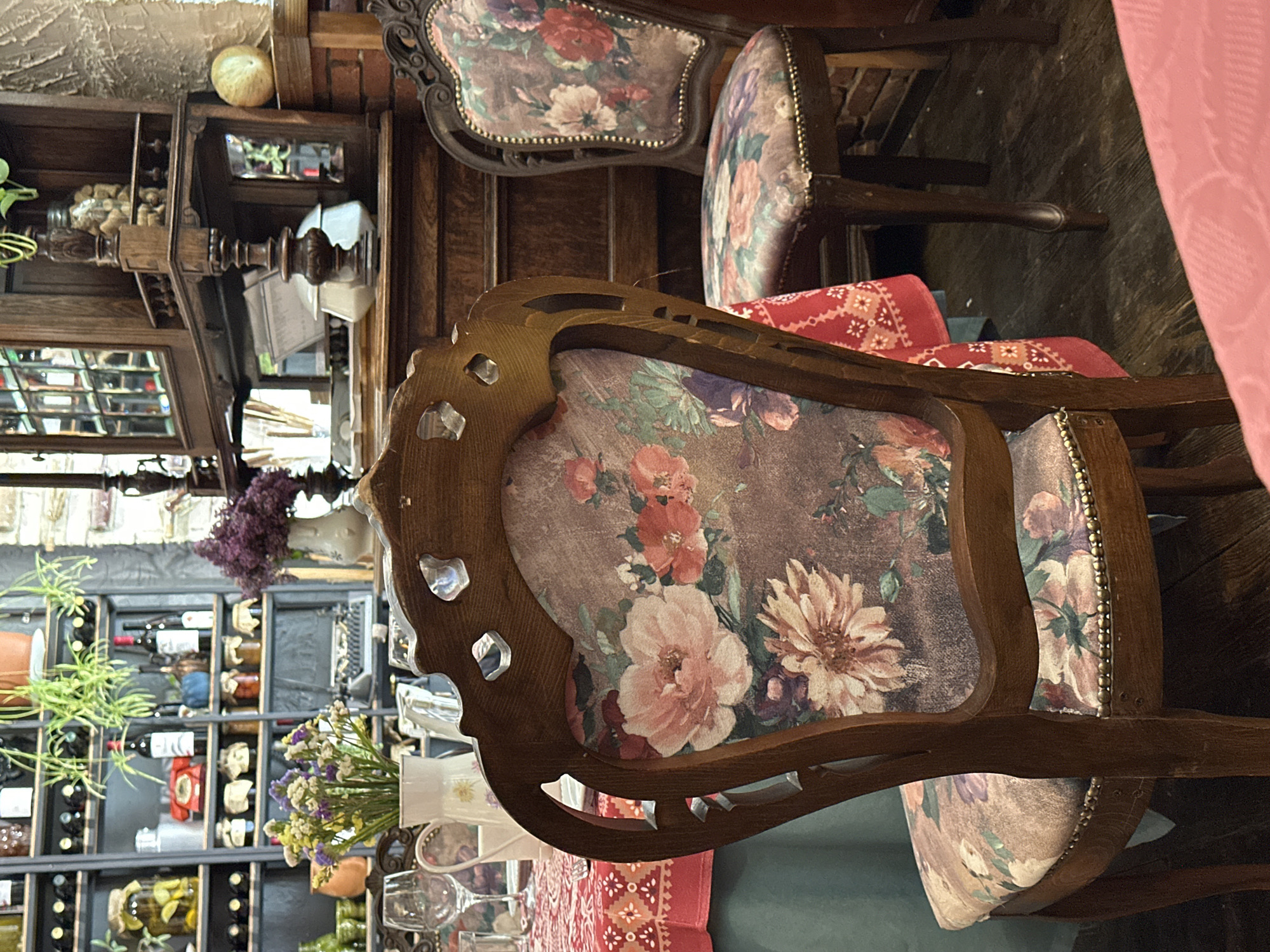 The image features a close-up view of two antique chairs with floral upholstery. The chairs are made of dark wood and have ornate designs. The foreground chair has a distinct, cut-out pattern along the top of the backrest. A red patterned cushion sits on the seat of the back chair. In the background, there is a wine rack filled with bottles and decorative objects. A floral arrangement is visible on a table near the chairs, with wine glasses placed on a red tablecloth. The scene appears to be set in a restaurant or dining area. Based on the context, this is most likely a dining location in the city of Tbilisi, Georgia.