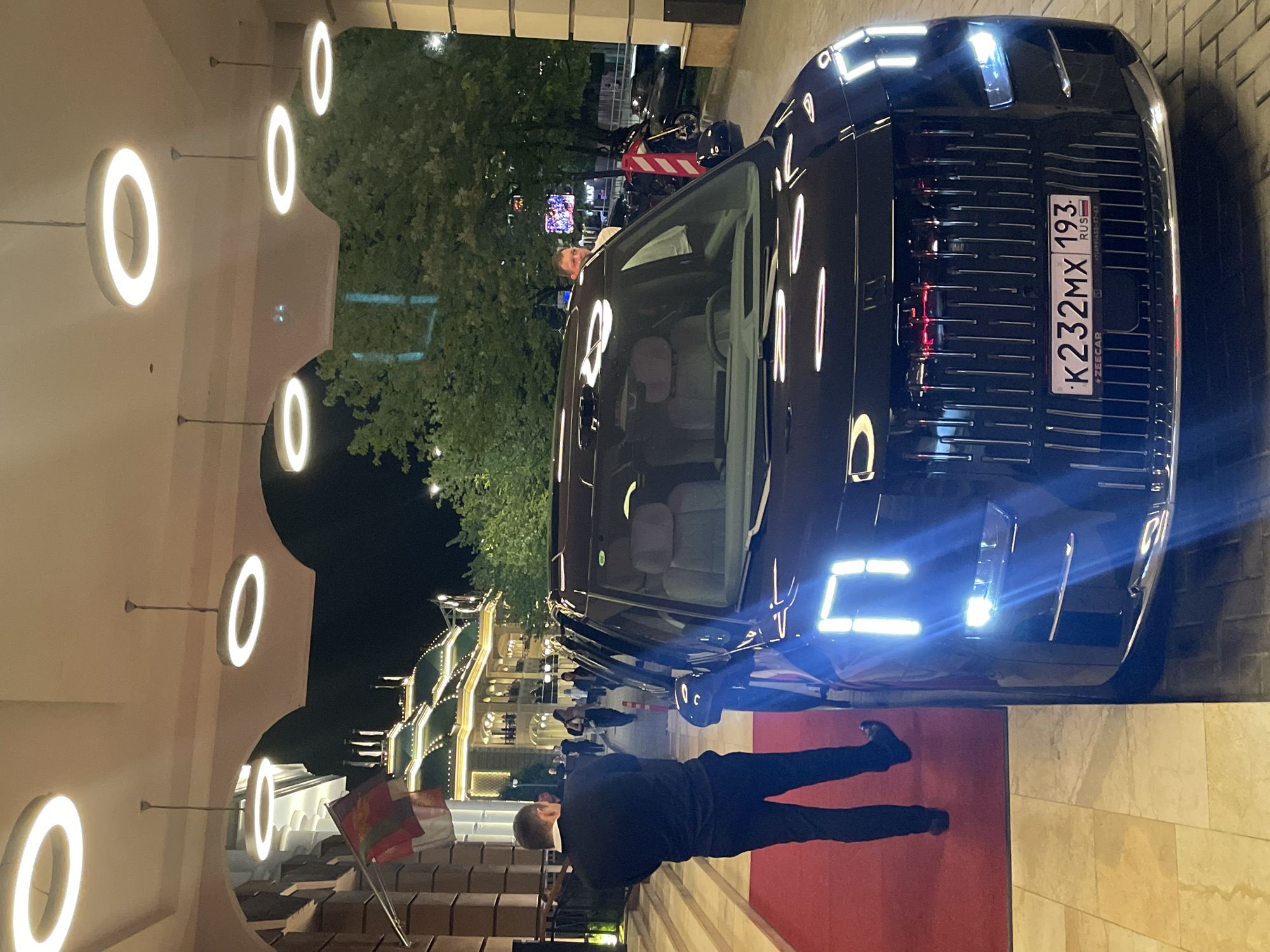 In the city of Estosadok, Russia, at night, a black car with distinctive LED headlights and a vertical grille is parked on a paved surface. A man in a dark suit and black pants stands on a red carpet, facing away from the camera, with his left hand raised. Behind him, several people are visible walking towards a brightly lit building with ornate architecture and a green roof. Circular pendant lights illuminate the covered walkway. Trees with dense foliage are present in the background. The car's license plate is partially visible and reads "K232MX 193".