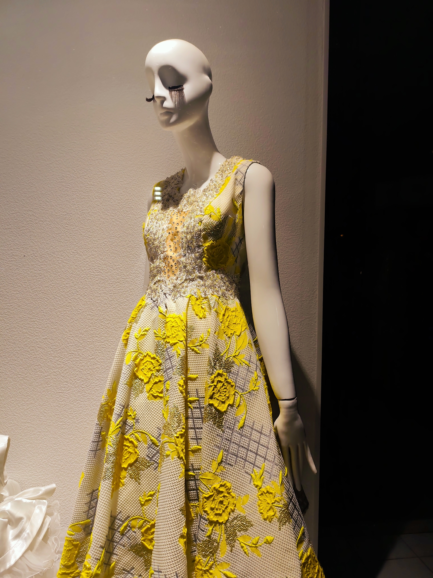 A mannequin wearing a formal yellow and white textured dress with floral embellishments and beaded details is displayed in a dimly lit store window in Fergana City, Uzbekistan. The mannequin has exaggerated eyelashes and appears to be crying or have makeup running down its face. The dress is sleeveless with a fitted bodice and a full skirt. The background is a textured beige wall and a dark doorway.