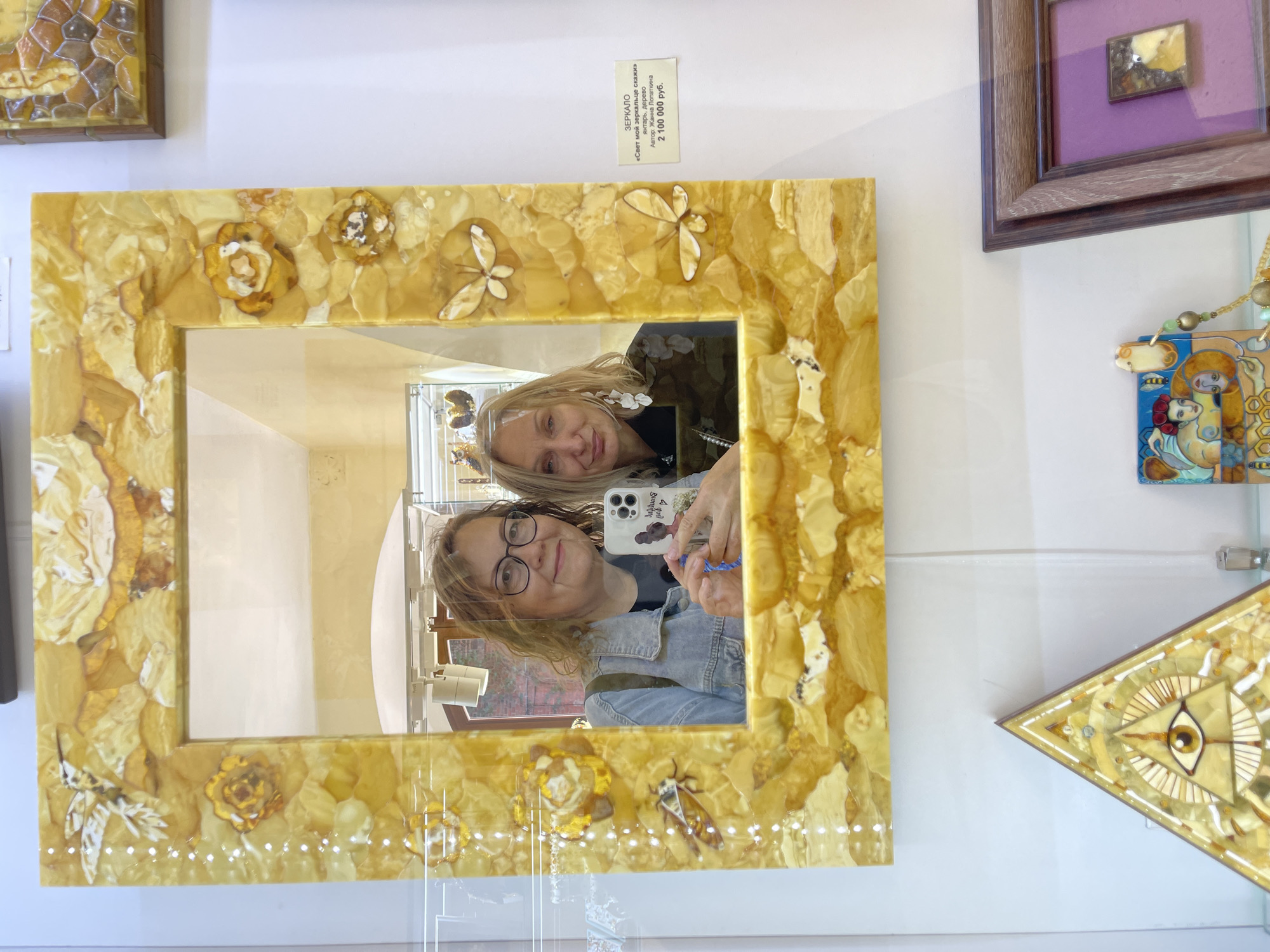 The image shows two women reflected in a mirror with an amber frame, likely in the Kaliningrad region of Russia, given the prevalence of amber artifacts there. The mirror is mounted on a white wall, surrounded by other framed artworks and displays. The woman in the foreground wears glasses and a denim jacket, holding a smartphone with a patterned case, as if taking a selfie. The second woman stands behind her and slightly to the side. The amber mirror frame is decorated with floral and insect motifs. A price tag is visible above the mirror, indicating a cost of 2,100,000 rubles.