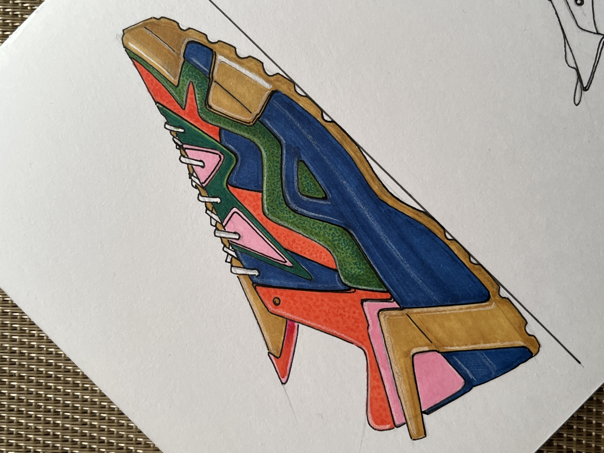 A colorful, stylized drawing of a shoe is positioned on a white surface. The shoe exhibits a variety of colors, including blue, green, orange, pink, and tan. The drawing is on a white surface that appears to be part of a paper pad, which is resting on a textured, tan surface.