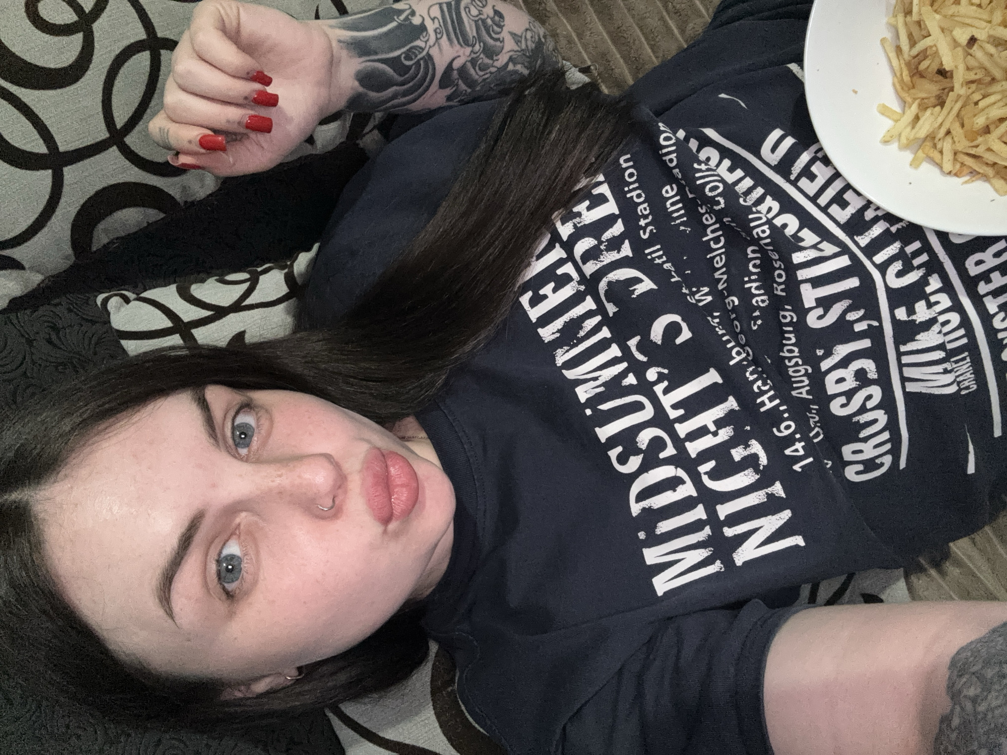 A woman is lying down, appearing to take a selfie. She has dark hair, fair skin, and red nail polish. She is wearing a black t-shirt with white text. A plate of french fries is visible in the upper right corner. The woman has tattoos on her arm. She is lying on a patterned couch. The OCR mentions "Latil Stadion, Wine, Augsburg, Rosenavi" - these appear to be text fragments from the t-shirt. There is no reference to Chornomorskoe, Ukraine in the image or OCR.
