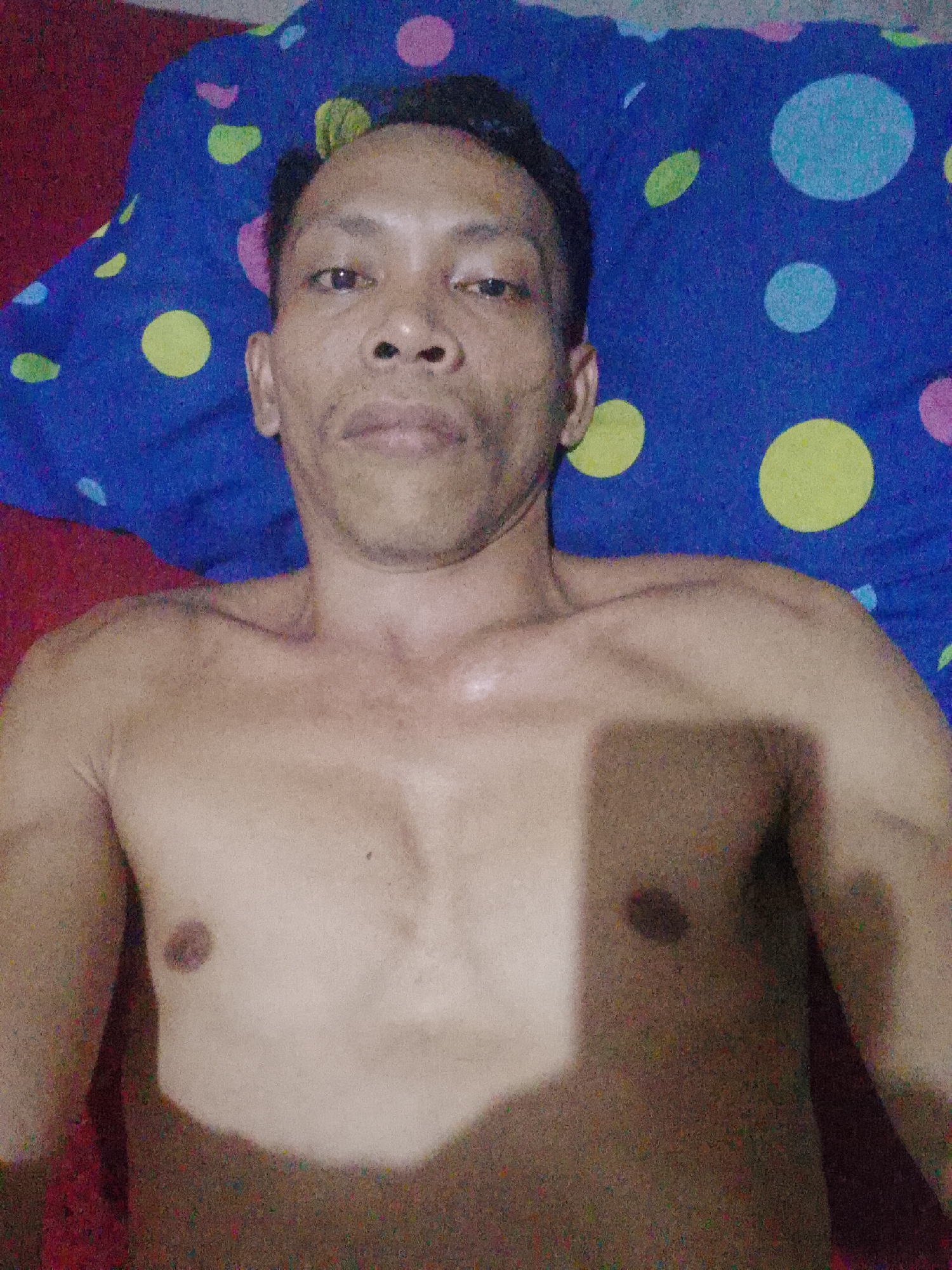 A shirtless man lies on a bed. The head of the bed is a blue and purple patterned fabric, while the bed itself is covered in a red fabric. The man appears to be in Bukit Ketok, Indonesia.