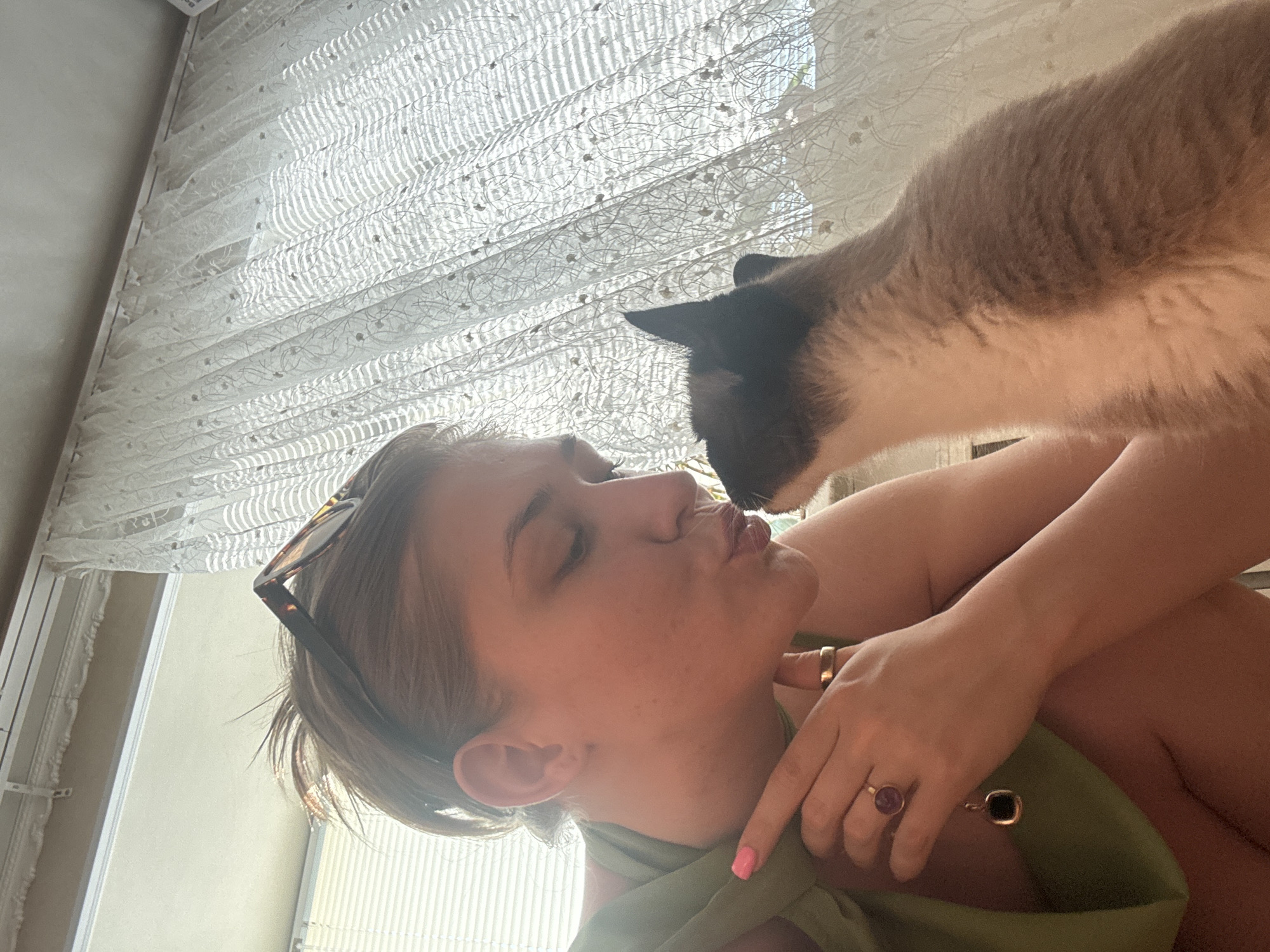 A young woman with sunglasses perched on her head and a green top is shown in profile, leaning towards a cat with Siamese markings. The woman is puckering her lips as if to kiss the cat, which is looking up at her. Her left hand is raised, with her index finger extended towards the cat. She is wearing a gold ring with a dark stone on her right ring finger, and a thin gold band on her left ring finger. A gold pendant with a black square stone hangs from her neck. The background features a window with blinds and a sheer white curtain with a floral pattern. The scene appears to be indoors, possibly in a residential setting in Ussurijsk, Russia.