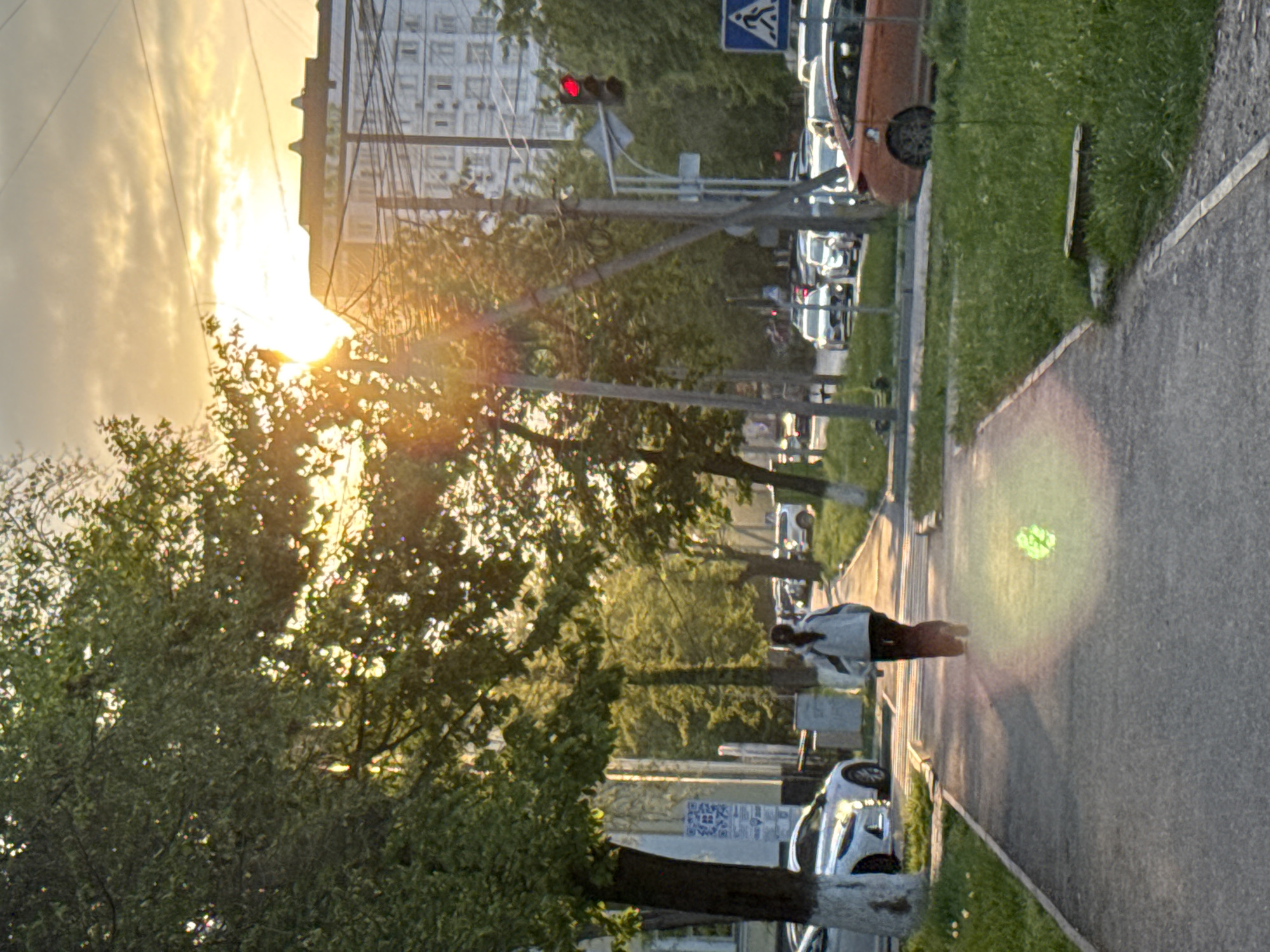 The image shows a street scene in Bishkek City, Kyrgyzstan. A person is walking on the sidewalk. Cars are parked along the street. Traffic lights are visible. A building is in the background. The sun is shining through the trees, creating lens flare.