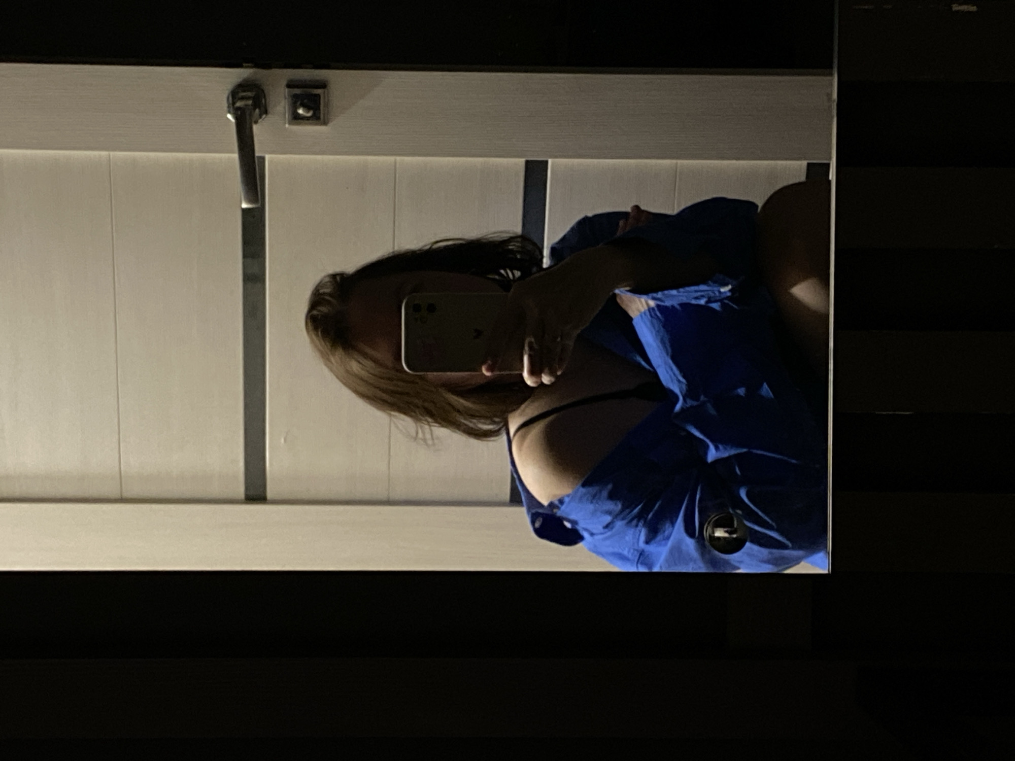 An indoor mirror selfie taken in Chaikovsky, Russia, features a woman obscured by her smartphone. She is shown from the chest up, with light brown hair and wearing a blue collared shirt or jacket, under which a black bra strap is visible. A circular logo, possibly a brand emblem, is present on her blue top.
The woman is holding a light-colored smartphone, likely an iPhone due to the visible multi-lens camera system and a partially visible Apple logo on its back. Her right hand is seen holding the phone, with fingers wrapped around it.
The background of the reflection reveals a room with white vertical wall paneling. In the upper right corner of the reflected scene (upper left in the provided image orientation), there are silver-toned fixtures: a door handle or latch and a small square hook or knob. The lighting in the scene is dim, suggesting it might be evening, night, or a poorly lit indoor environment. The setting appears to be a private interior space, possibly a bathroom or changing room. No other discernible text is visible besides the faint Apple logo on the phone.