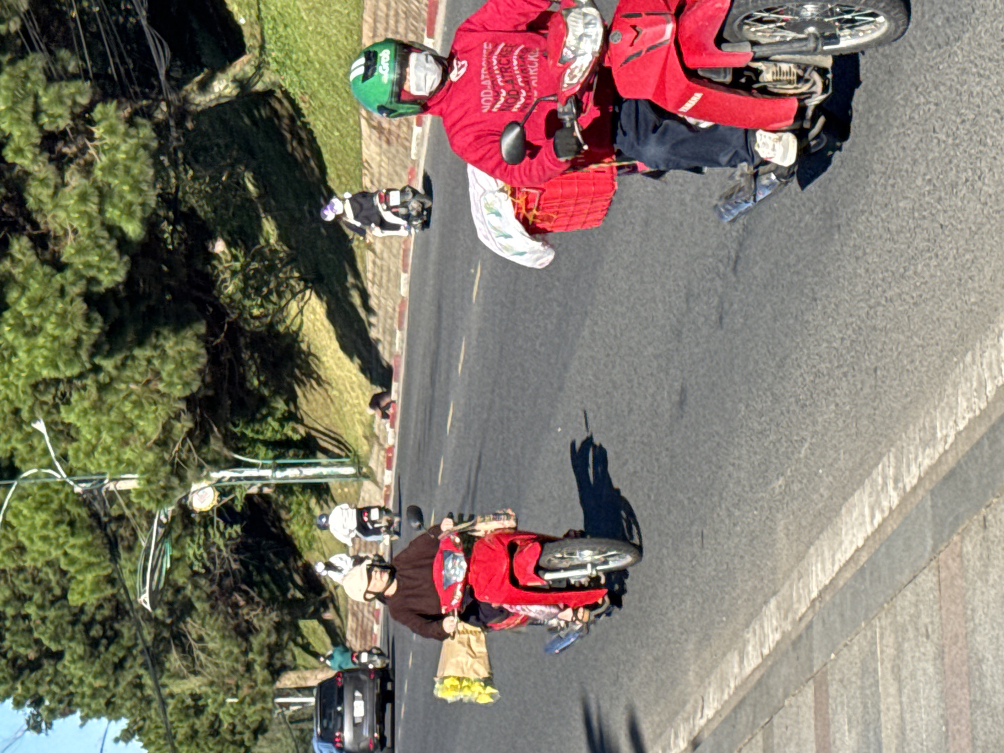 In Da Lat, Vietnam, a woman wearing a light-colored helmet and a dark brown shirt rides a red motorcycle on a paved road. She is carrying a large bouquet of yellow flowers wrapped in brown paper to her left. In front of her, the motorcycle has a clear plastic bag attached to the handlebars. Behind her, a red basket is mounted on the rear of the motorcycle, partially covered by a white cloth.
In the foreground, to the right, a person wearing a green helmet with "Grab" branding, a red jacket, black pants, and a white face mask rides a red motorcycle. This person also has a red basket mounted on the rear, with a white cloth covering its contents. The person is wearing black gloves.
Further in the background, several other individuals on motorcycles are visible. One person in a white helmet and black clothing is riding a dark-colored motorcycle. Another person in a white helmet and purple jacket is also on a dark motorcycle. A person in a white helmet and a green top is on a dark motorcycle. To the far left, a dark gray car is parked. The road is lined with trees and green slopes, indicating an outdoor urban or suburban environment, consistent with Da Lat's landscape. The lighting suggests it is daytime, with shadows cast by the trees and vehicles.