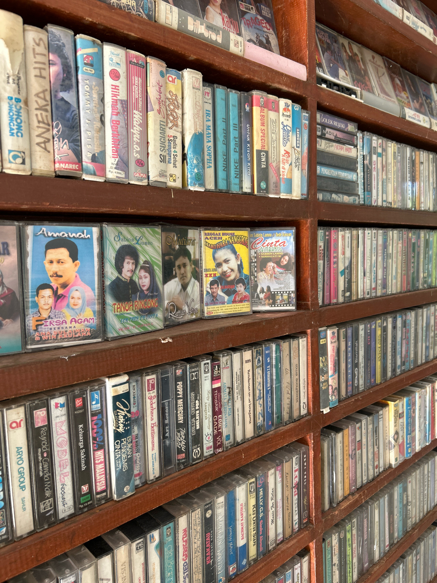 The image shows a close-up view of a wooden bookshelf filled with cassette tapes. The tapes are organized in rows and appear to be primarily music albums. The titles and artwork on the cassette cases are varied, with some featuring portraits of musicians and others displaying graphic designs. Some of the visible text suggests the content is in Indonesian, with titles like "Aneka Hits," "Amanah," and "Sabirin Lamno." The bookshelf itself is a dark wood with visible grain, and the overall lighting is warm and somewhat dim, suggesting an indoor setting. The presence of numerous cassette tapes indicates a collection, likely for personal use or sale. The image is focused on the tapes, with the background being the shelves and a hint of more shelves beyond. There are no people clearly visible in the image, and no actions are taking place. The context provided about Sabang, Indonesia, implies this collection is located in a shop or private residence within that city, reflecting a possible cultural or historical aspect of music distribution in the region.