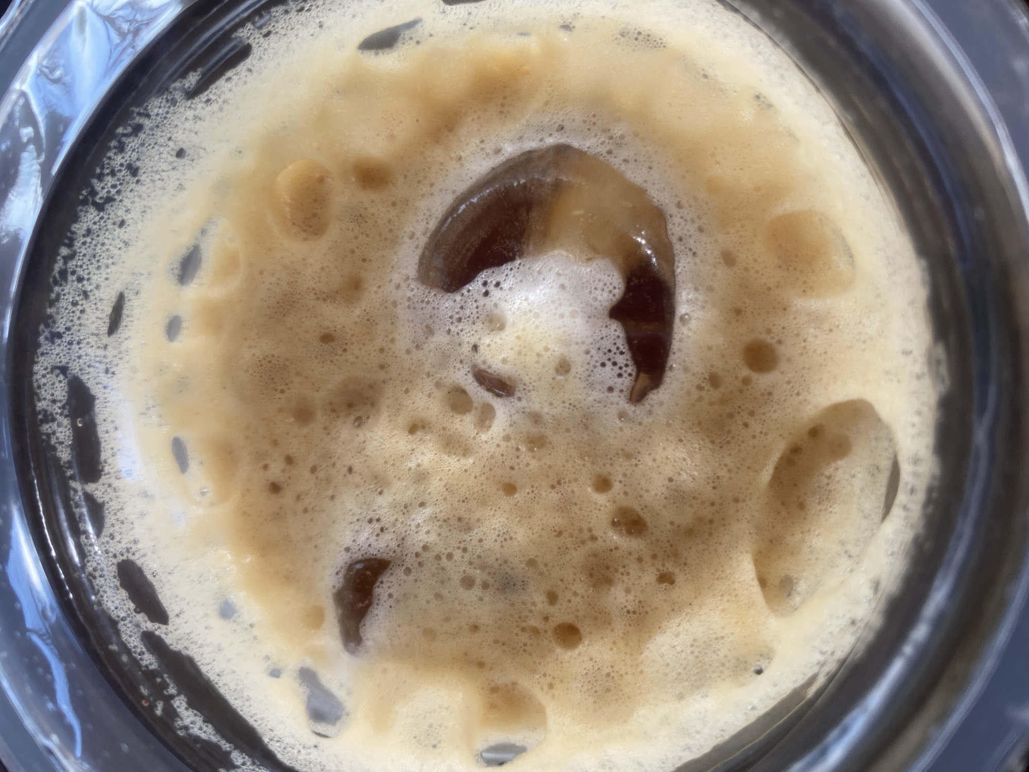 A close-up, top-down view shows an iced coffee beverage in a clear glass. The coffee is topped with a thick layer of foam, creating a creamy, light brown surface dotted with small bubbles. Several dark brown, irregularly shaped ice cubes are partially submerged in the foamy mixture. The reflections in the clear glass suggest an outdoor setting, possibly a cafe or a patio, with blue sky and sunlight visible. The overall impression is a refreshing, cold drink. There is no information available about the location in the city of Lhokseumawe, Indonesia.