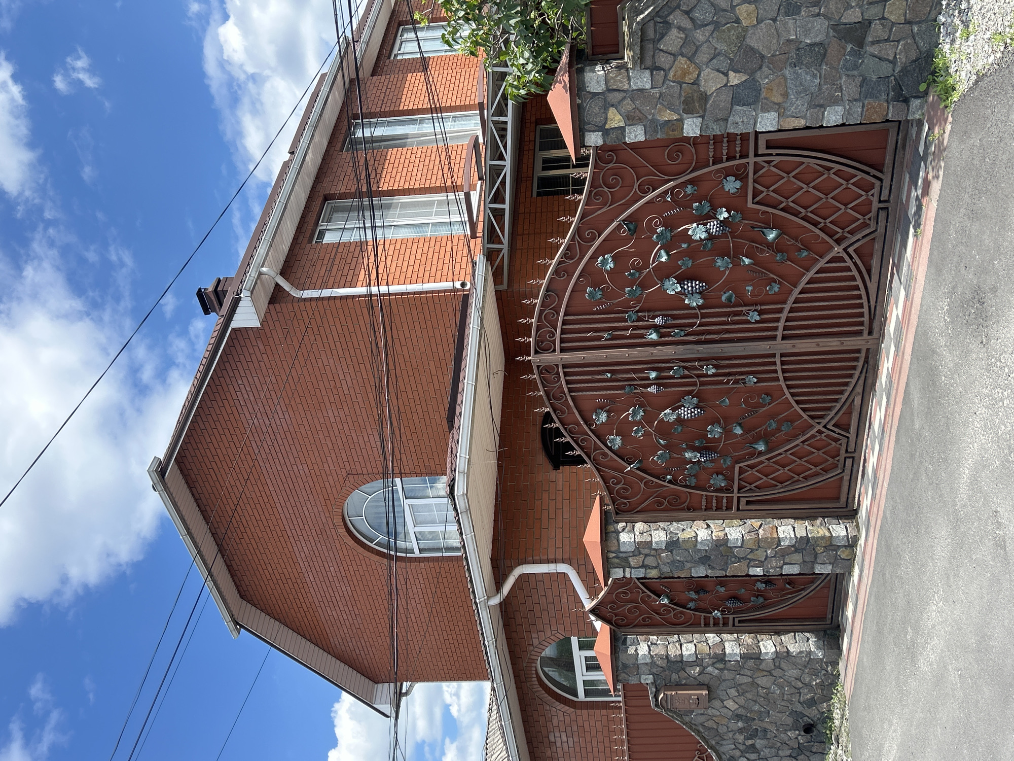 The media file depicts the exterior of a brick house with an ornate, dark brown metal gate featuring a grape vine motif. The gate is partially open, revealing a glimpse of the property beyond. The house has multiple windows, some of which are arched and feature white frames. The exterior walls are constructed from red brick, and the roof appears to be tiled. Adjacent to the gate and the house's facade is a wall made of rough-hewn stones. The scene is set against a bright blue sky with scattered white clouds, suggesting a clear, sunny day. Power lines are visible running across the sky and near the house, indicating an urban or suburban setting. The image is captured from a low angle, looking upwards towards the house and sky, giving a sense of scale and height. The overall impression is one of a well-maintained, possibly older, residential property in Bila Tserkva, Ukraine. No people are visible in the media. The time of day appears to be daytime, likely late morning or early afternoon, given the bright sunlight.