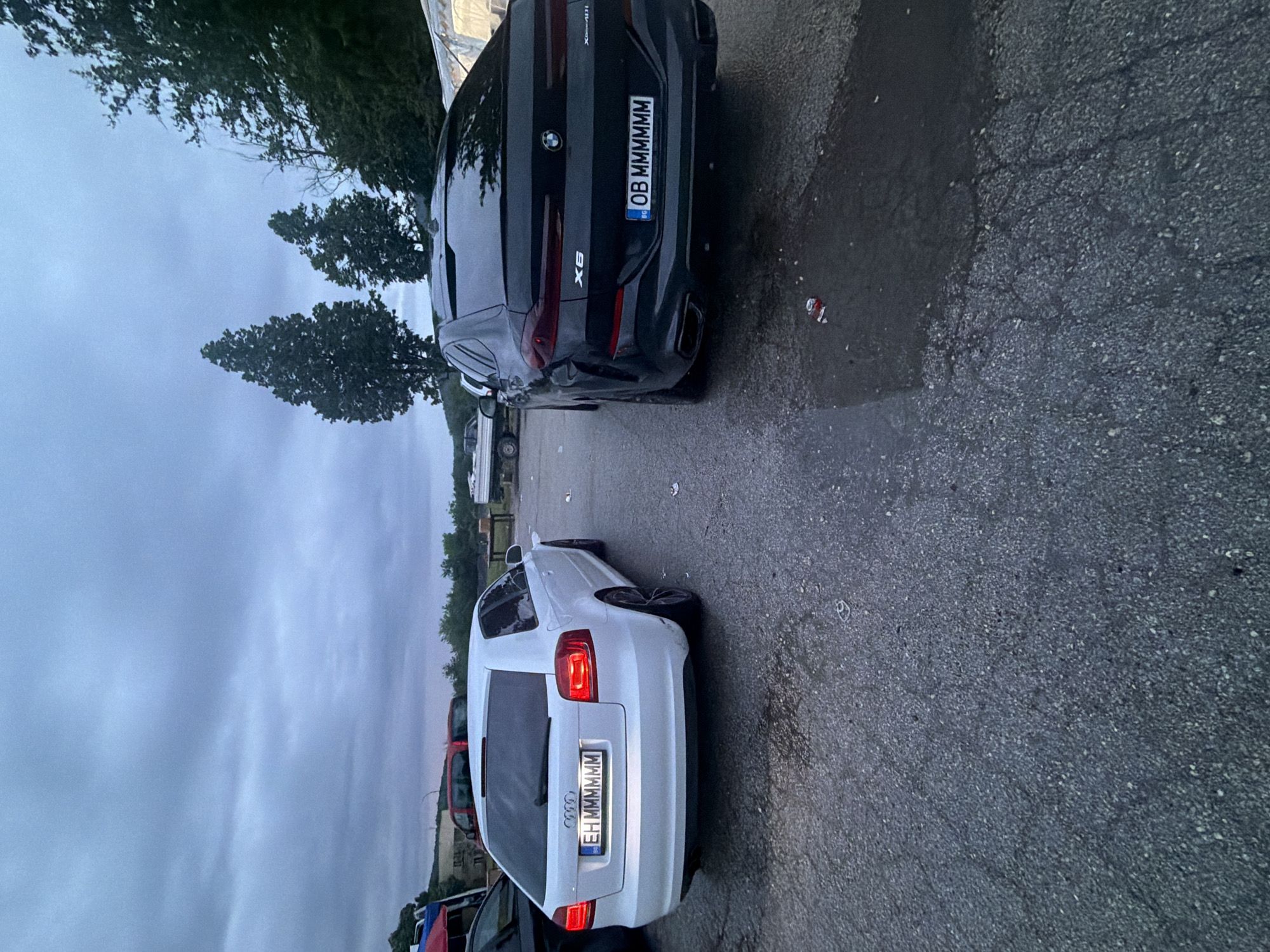 A black BMW X6 and a white Audi A3 are parked side-by-side in a parking lot. The BMW's license plate reads "OB MMMMMM" and the Audi's reads "EH MMMMMM". The photo was taken in Zlatna Panega, Bulgaria.