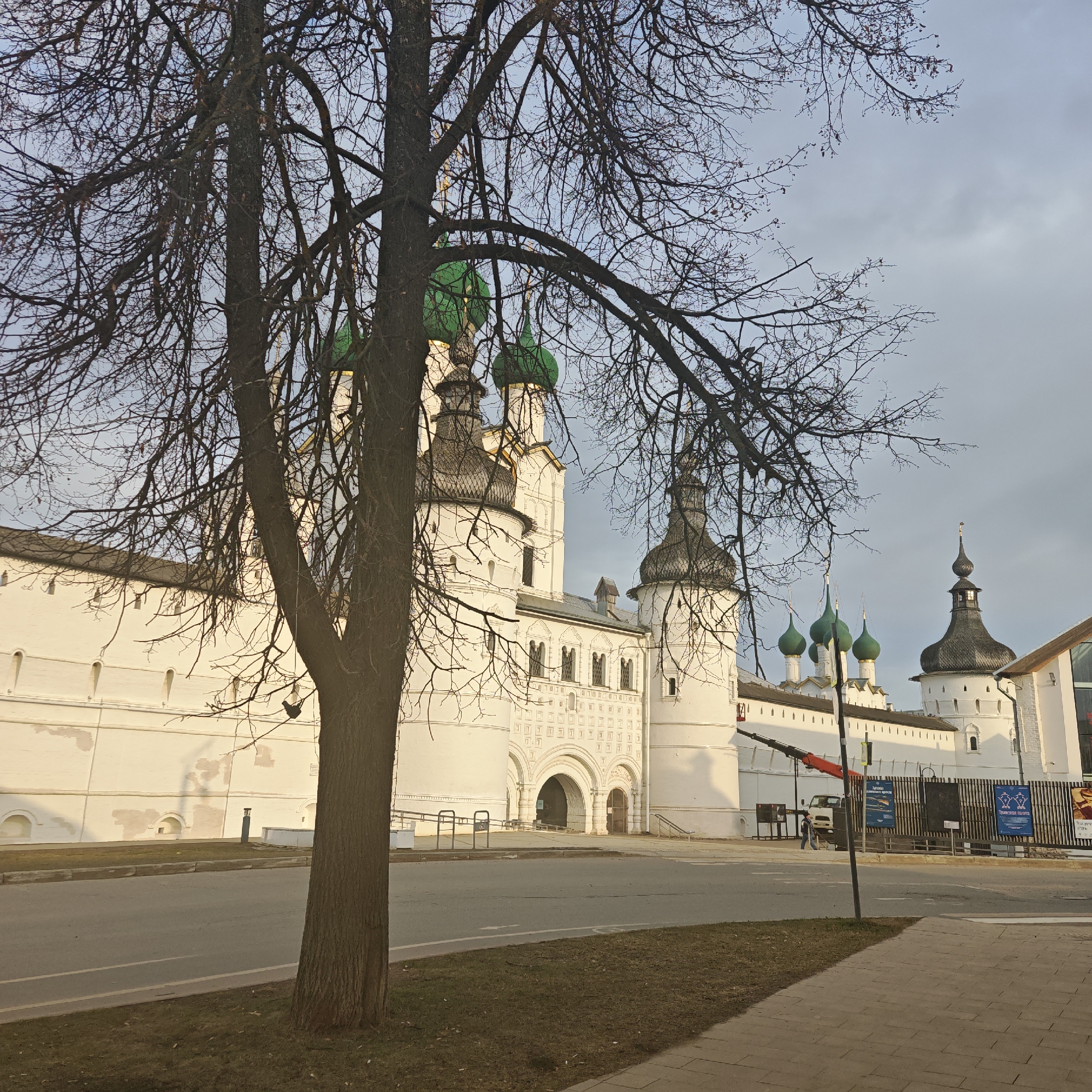 The image shows a street scene in Rostov, Russia, with a prominent white-walled structure featuring green domes, identified as the Rostov Kremlin. A bare tree stands in the foreground, partially obscuring the view. A wide street runs horizontally through the scene, dividing a grassy area on the left from a paved sidewalk on the right. In the background, there is construction equipment, and a fence with signage. A person is visible in the distance near the construction. The sky is overcast.