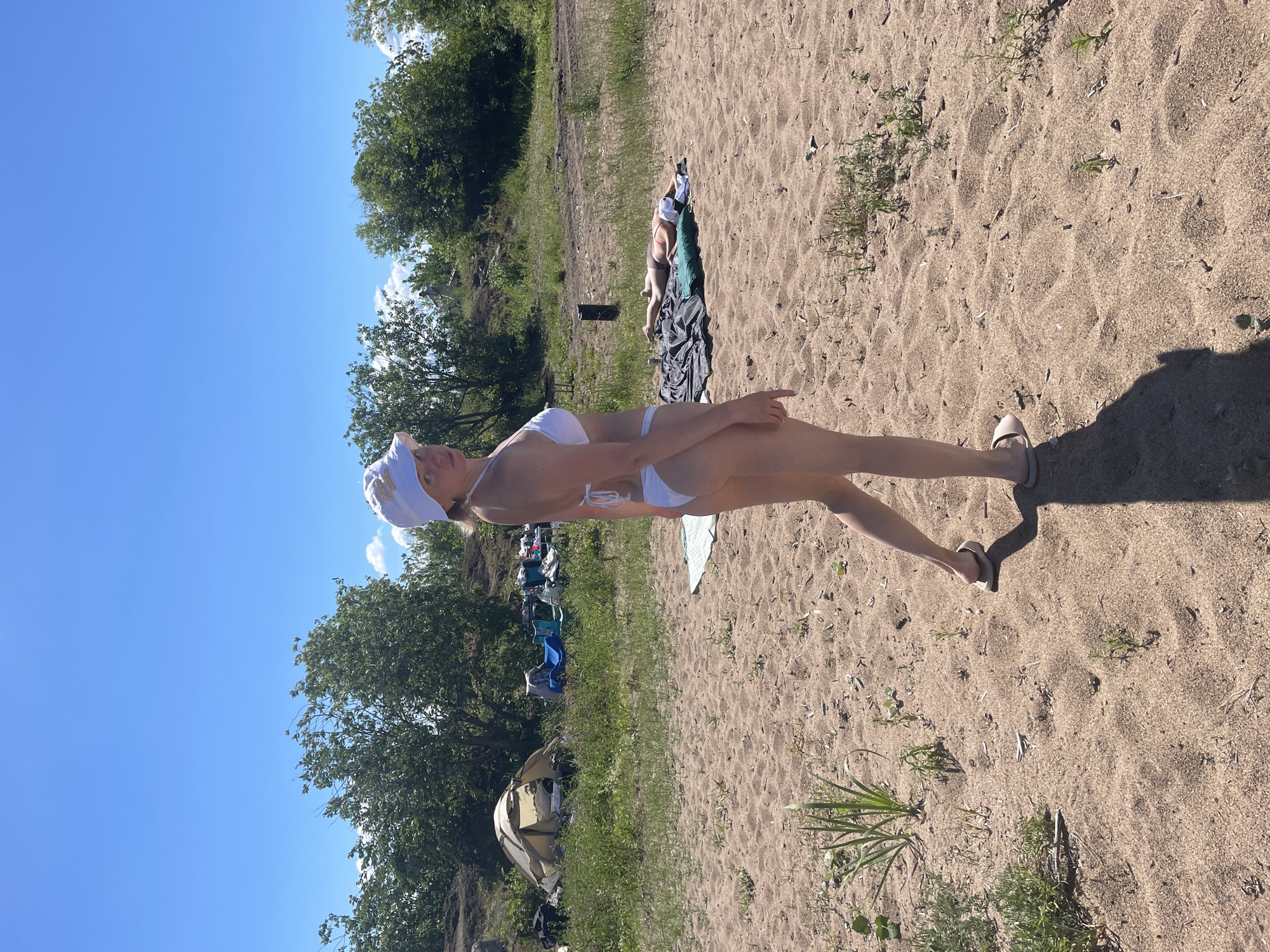 This media file depicts a sunny day at a sandy beach near Belozersk, Russia. The main subject is a woman wearing a white bikini and a white headscarf, posing on the sand. Her body is oriented sideways to the camera. In the background, another person is lying on a dark blanket with a green pillow. Further back, several camping chairs and a tent are visible near a grassy area with trees. The sky is a clear, bright blue, suggesting a warm and pleasant day. The sand is textured with ripples and scattered vegetation. The presence of camping gear and the natural setting indicate a recreational outing. No visible text is present in the image.