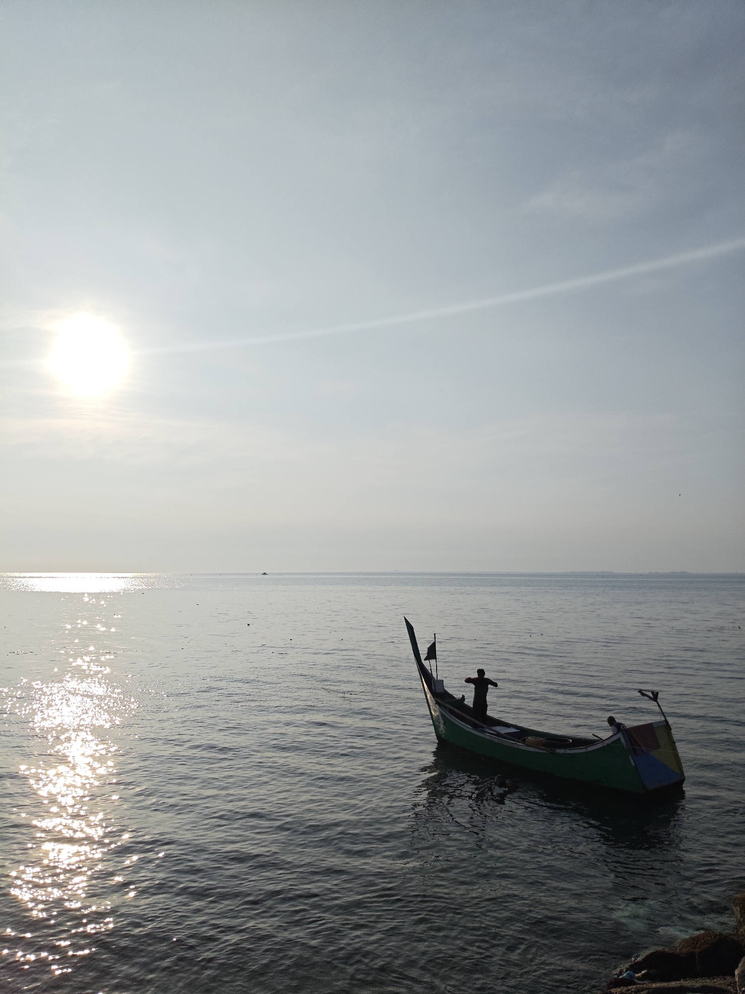 A serene coastal scene in Lhokseumawe, Indonesia. A lone, traditional green fishing boat is anchored near the shore. An individual stands on the boat, appearing to be preparing for fishing. The water is calm with shimmering sunlight reflecting off its surface. In the distance, another small boat can be seen. The sky is bright and clear with the sun shining. Some rocks are visible at the very bottom of the shot, suggesting the proximity of the shoreline.