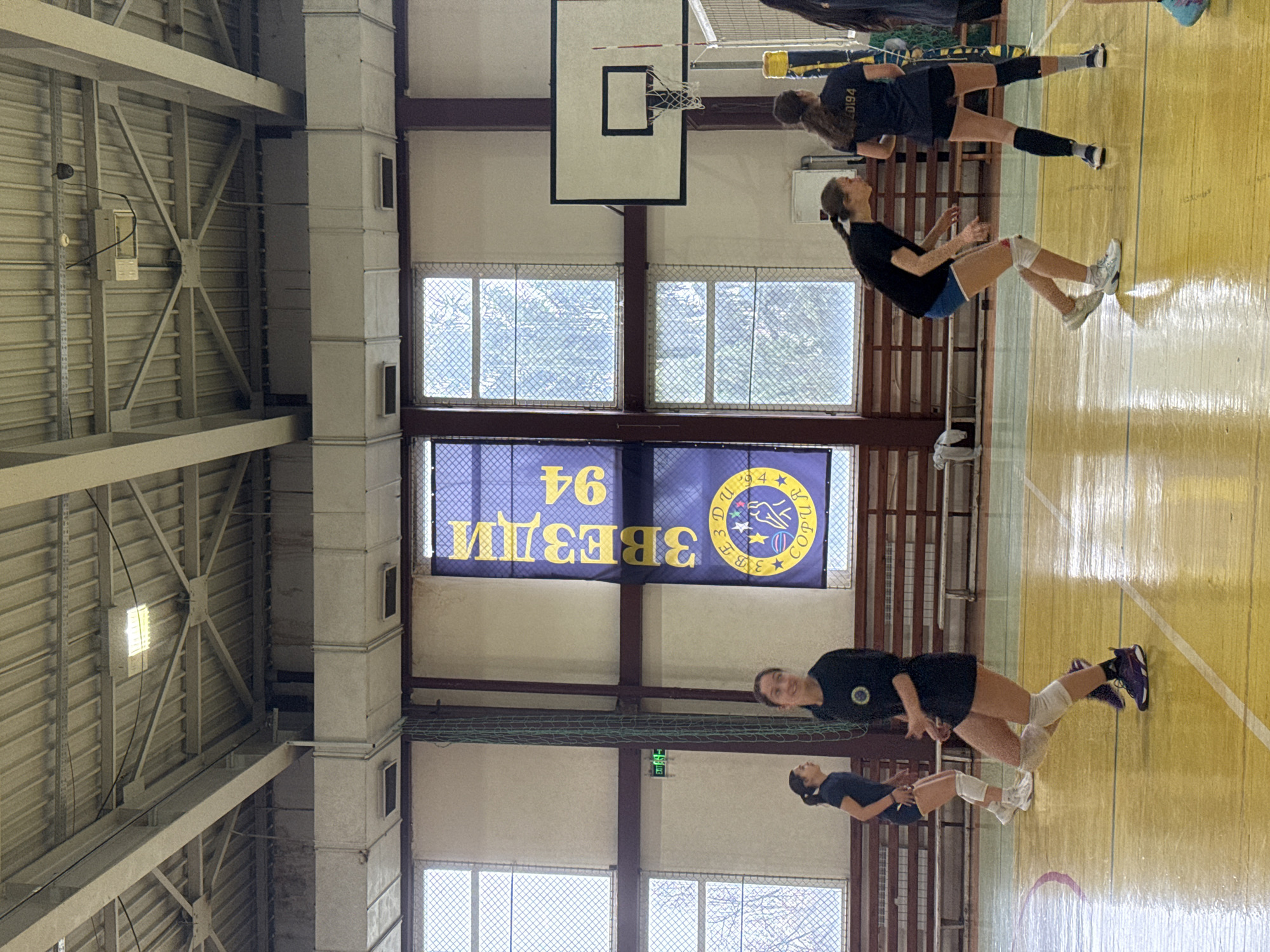 The scene depicts an indoor sports gymnasium in Sofia, Bulgaria, identifiable by the prominent blue and yellow banner on the back wall displaying "3 ЗВЕЗДИ 94" (3 STARS 94) and "СОФИЯ" (SOFIA) in Cyrillic, along with a club logo.
Six female individuals, likely young adults or adolescents, are present on a polished wooden court with visible green and red boundary lines, engaged in what appears to be a volleyball practice or game. All subjects are dressed in athletic attire, including dark t-shirts, shorts, and knee pads.
In the foreground, slightly left of center, one player in a dark top, dark shorts, white knee pads, and purple athletic shoes is in a ready stance, looking towards the left. Behind her and to the left, another player in similar attire and white knee pads is also in a ready stance. To the right, a third player, wearing a dark top, blue shorts, white knee pads, and white athletic shoes, is crouched low. Further right, near the volleyball net, a player with her back partially to the camera wears a dark top with "20194" printed on it, dark shorts, and black knee pads. Two other players are partially visible on the far right, adjacent to the net.
The environment features a high industrial ceiling composed of corrugated metal sheets supported by exposed metal trusses and beams, with several rectangular and square artificial light fixtures. White rectangular ventilation ducts run horizontally across the upper wall section, above a series of large, multi-paned windows. These windows provide natural light and are partially obscured by a wire mesh covering; bare tree branches are visible through them. A set of light brown wooden spectator benches lines the far wall. A basketball hoop with a white backboard and black rim is positioned on the right side of the court, behind the volleyball net, which has a yellow and blue top band. The court's floor reflects the overhead lighting.