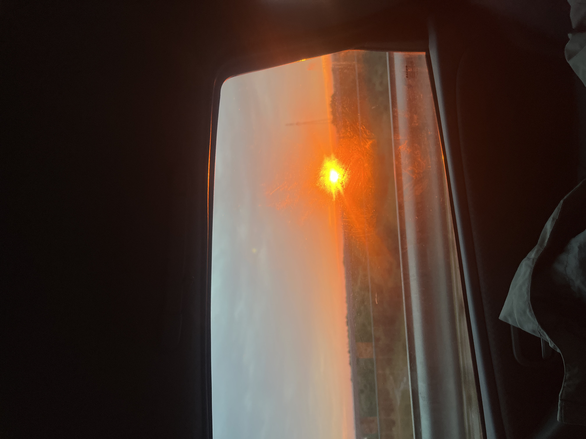The image is a photograph taken from inside a vehicle, looking out a window at a sunset. The sun is a bright orange orb low in the sky, casting a warm glow across the horizon and illuminating the clouds with shades of orange and pink. The landscape outside appears to be rural, with trees and some structures visible. The window itself is partially obscured by streaks and smudges, suggesting it may be dirty or wet. The interior of the vehicle is dark, with the lower portion of the frame showing part of a car door and a dark fabric item, possibly a blanket or bag. The overall impression is one of a scenic sunset viewed during travel, with the specific location being within the Азяранскі сельскі Савет in Belarus.