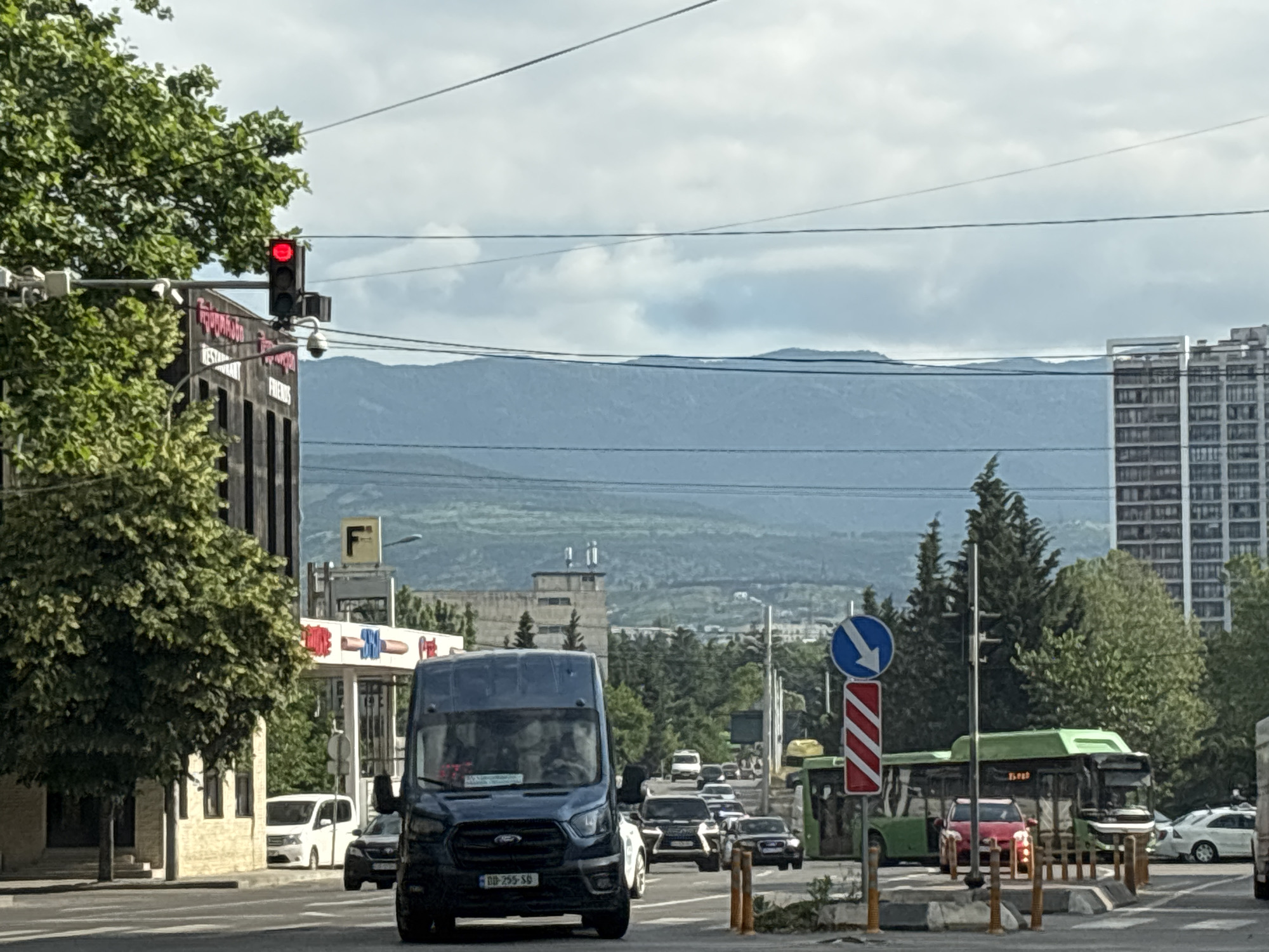 The image captures a street scene in Tbilisi, Georgia, on a cloudy day. A dark blue Ford Transit van is prominently featured in the foreground, stopped at a red traffic light. The van has a Georgian license plate that reads "BD 755 SB". Multiple passengers are visible inside the van, appearing to be looking forward. To the left of the van, a street tree with lush green foliage partially obscures a building with a sign that reads "RESTAURANT FRIENDS" in Cyrillic and Latin script. Further left, a red traffic light is illuminated, indicating a stop. Behind the van, other vehicles, including cars and a white van, are also stopped or moving in traffic. A green bus is visible on the right side of the road, with a red car in front of it. In the background, a tall, modern residential building with many windows stands out on the right, while a lower, industrial-looking building with a tower is visible in the center distance. Rolling, blue-hued mountains form the backdrop under a sky filled with soft, white clouds. The scene is characterized by urban infrastructure, including traffic lights, street signs, and power lines.