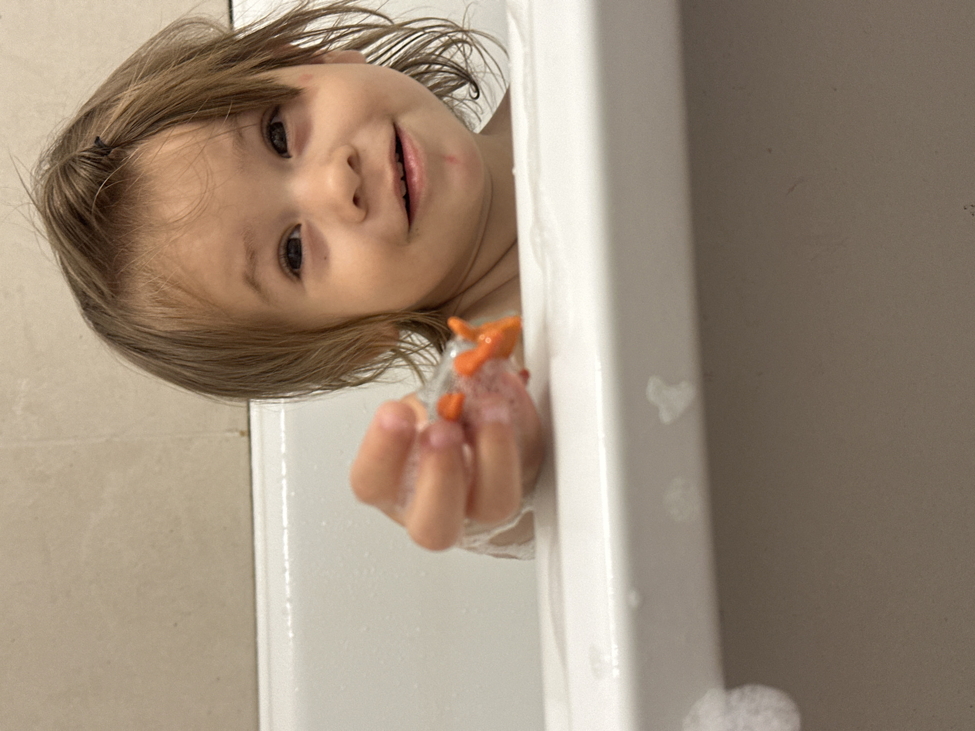 A child is in a white bathtub, holding an orange starfish toy. The child has light brown hair and dark eyes. Bubbles are present in the water. The bathroom has beige tiles. The location is not discernible as Kharnavy, Russia from this image.