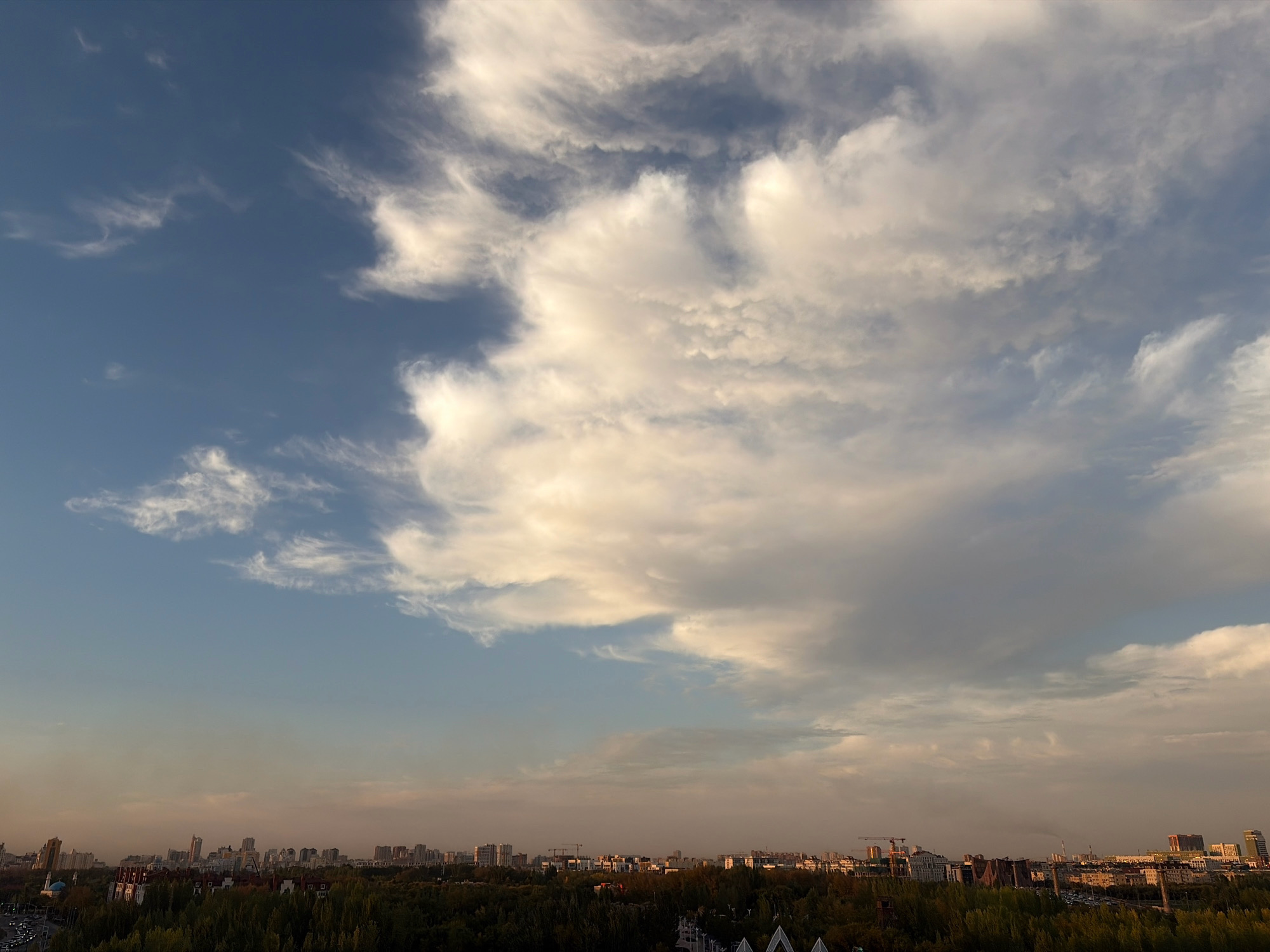 This image depicts an aerial view of the city of Astana, Kazakhstan, at sunset. The sky is a dramatic canvas of contrasting blue and white clouds, with large, fluffy formations dominating the upper right portion of the frame and wispy cirrus clouds stretching across the horizon. The lower half of the image showcases the urban landscape, characterized by a dense line of trees forming a natural barrier between the viewer and the city's skyline. Beyond the trees, a multitude of buildings of varying heights and architectural styles are visible, with some taller structures and residential blocks defining the cityscape. The overall impression is one of a bustling metropolis bathed in the warm, fading light of the sun.
