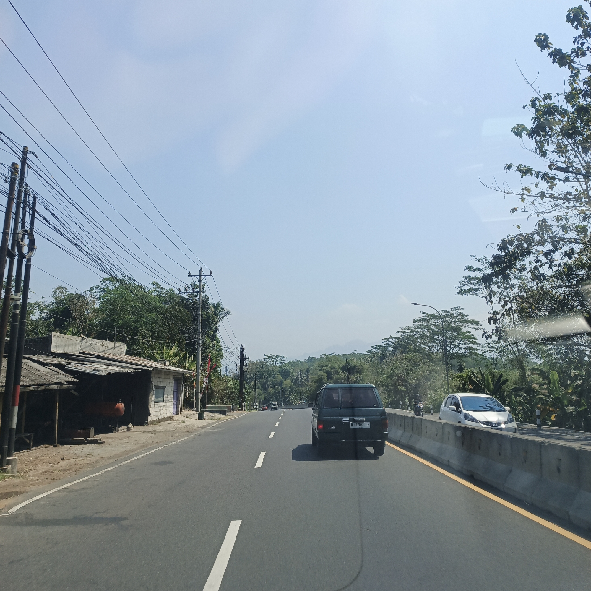 The image depicts a view from a vehicle traveling on a road in Secang, Indonesia, likely on a sunny day with a clear sky. On the left side of the road, there is a small, somewhat dilapidated building with a corrugated metal roof. Adjacent to the building are utility poles with a dense network of electrical wires overhead. The road itself is asphalt, with white dashed lines indicating lanes.
In the foreground and mid-ground, a dark green SUV is traveling ahead, its license plate readable as "H 1286 GF". Further ahead on the right side of the road, a white compact car is visible, and beyond it, a concrete barrier separates the lanes. The roadside is lined with lush green trees and vegetation, characteristic of a tropical environment. In the distance, rolling hills are visible under the hazy sky. A single streetlight is also present on the right side of the road. The overall scene suggests a typical rural or suburban road in Indonesia.