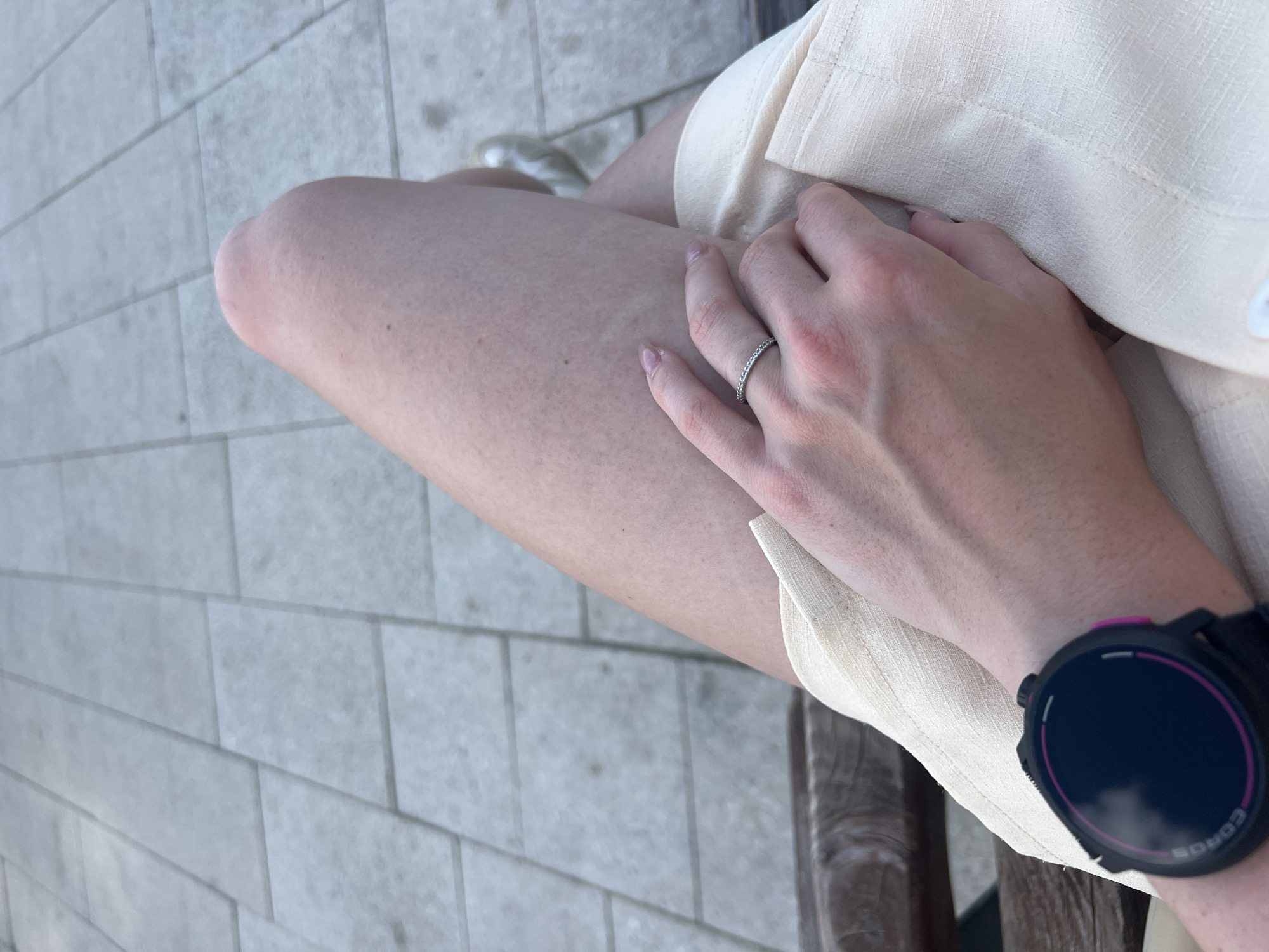 A person is sitting outdoors on what appears to be a bench. The person is wearing light beige shorts, a silver ring on their left ring finger, and a black and purple COROS watch on their left wrist. The person's left hand is resting on their leg. The ground is paved with rectangular gray blocks. The location is likely outdoors, but the specific place in Chaikovsky, Russia, is not evident from the image.