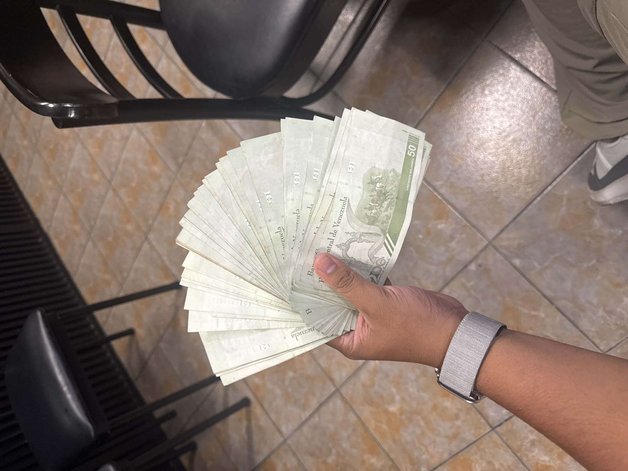 A person is holding a large stack of Venezuelan currency, specifically 50 Bolívar banknotes. The person's hand and wrist, adorned with a grey watch band, are visible. The background includes a black metal chair, light brown tiled floor, and part of a person's leg wearing grey pants and a white sneaker. The location is inferred to be in Parroquia Ocumare, Venezuela based on the currency.