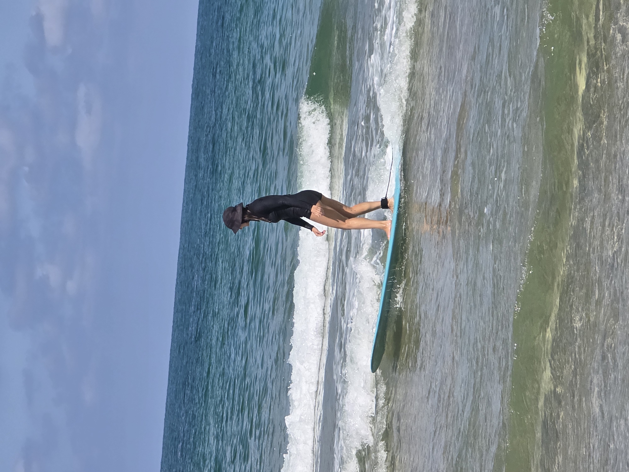 A woman is surfing on a turquoise surfboard in the ocean. The image is taken at an angle, so the sky appears on the left, the ocean on the top, and the shoreline on the right. The woman is wearing a dark-colored wetsuit and a hat. The ocean water is turquoise with white foam from the waves. The location could be the city of Tel-Aviv, Israel, but this is not definitively verifiable from the image.