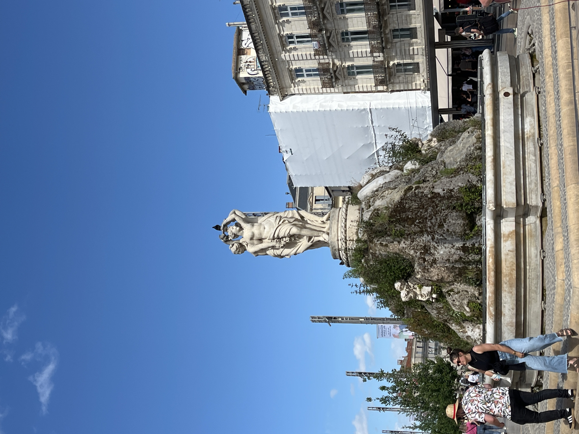 The image depicts the Fountain of the Three Graces in Montpellier, France, set against a clear blue sky. The statue is centered in the frame, showing the three figures in a classical pose. The fountain is constructed of marble, with moss and greenery growing on its base. To the right of the fountain, pedestrians are visible, including a woman wearing jeans and a black top. In the background, a building is covered with a white tarp.