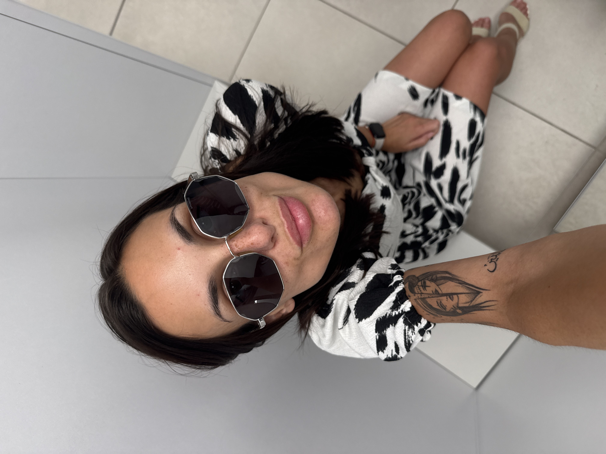 A young woman is captured in a high-angle shot, lying on her back and looking directly at the camera with a subtle, relaxed smile. Her head is slightly elevated. She is wearing a short-sleeved dress featuring a striking black and white abstract or animal print pattern.
Her dark hair is visible around her head. She sports dark, hexagonal-framed sunglasses, with the brand "Polaroid" faintly visible on the top right frame. A black-strapped smartwatch is worn on her left wrist. On her right forearm, two black tattoos are visible: a stylized depiction of a face or figure with flowing hair, and a small Om symbol positioned above it. Her bare legs are bent, and she wears light-colored, possibly cream or beige, open-toed sandals.
The setting appears to be indoors, with the woman resting on a light-colored, possibly off-white, tiled floor. The background consists of plain, light grey or white walls or surfaces, creating a clean, minimalist environment. The lighting is bright and even, indicating an indoor setting during the daytime.
The image is a casual, relaxed portrait, likely a selfie, offering a unique perspective of the subject. The location is Pleven, Bulgaria.