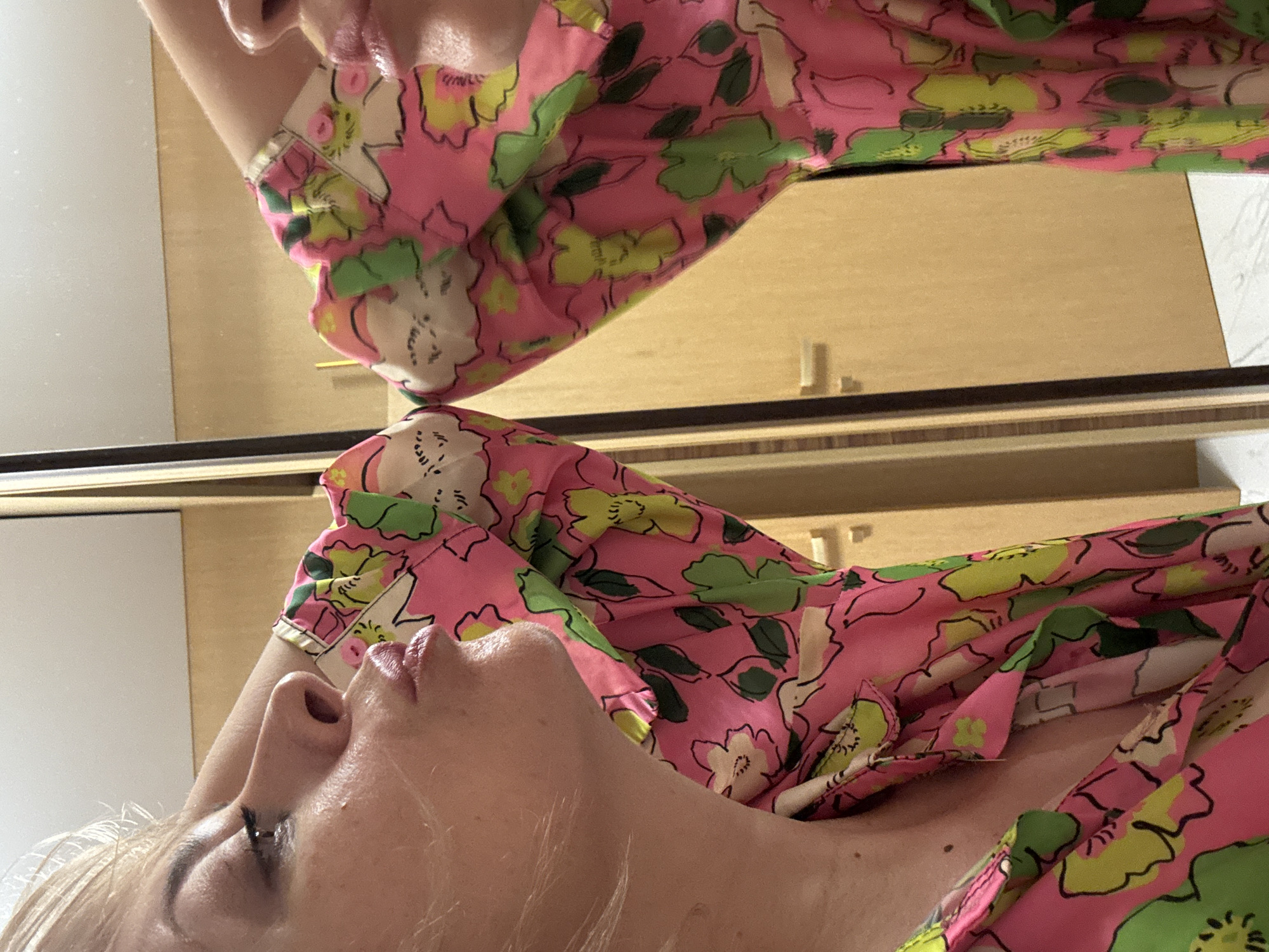 An adult female with blonde hair is captured in a profile view, leaning forward towards a mirror. Her lips are puckered, as if to kiss her reflection. She is wearing a pink garment adorned with a bold floral pattern featuring green leaves and yellow and white flowers. The background includes a reflective surface, likely a mirror, revealing a portion of her body and the same patterned garment. To the right of the mirror, a wooden door with a handle is visible. The scene appears to be indoors, possibly in a room with light-colored walls and flooring. The image does not provide any information that would allow for a definitive determination of its location in Sochi, Russia.