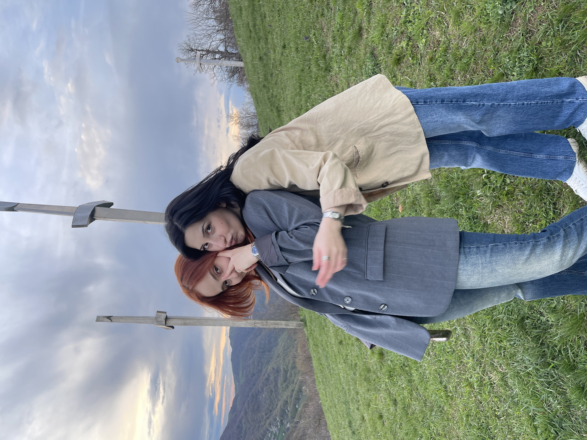 Two women are posing for a photo in what appears to be a grassy field. One woman has black hair and is wearing a gray blazer and jeans. She has her arm around the other woman and is lightly touching her face. The second woman has red hair and is wearing a tan jacket and jeans. They appear to be standing on a hillside overlooking a valley with hills in the distance. In the upper-left corner of the image, there is a gray metal post or structure. The sky is cloudy with hints of sunlight. The location is Shamta, Georgia.