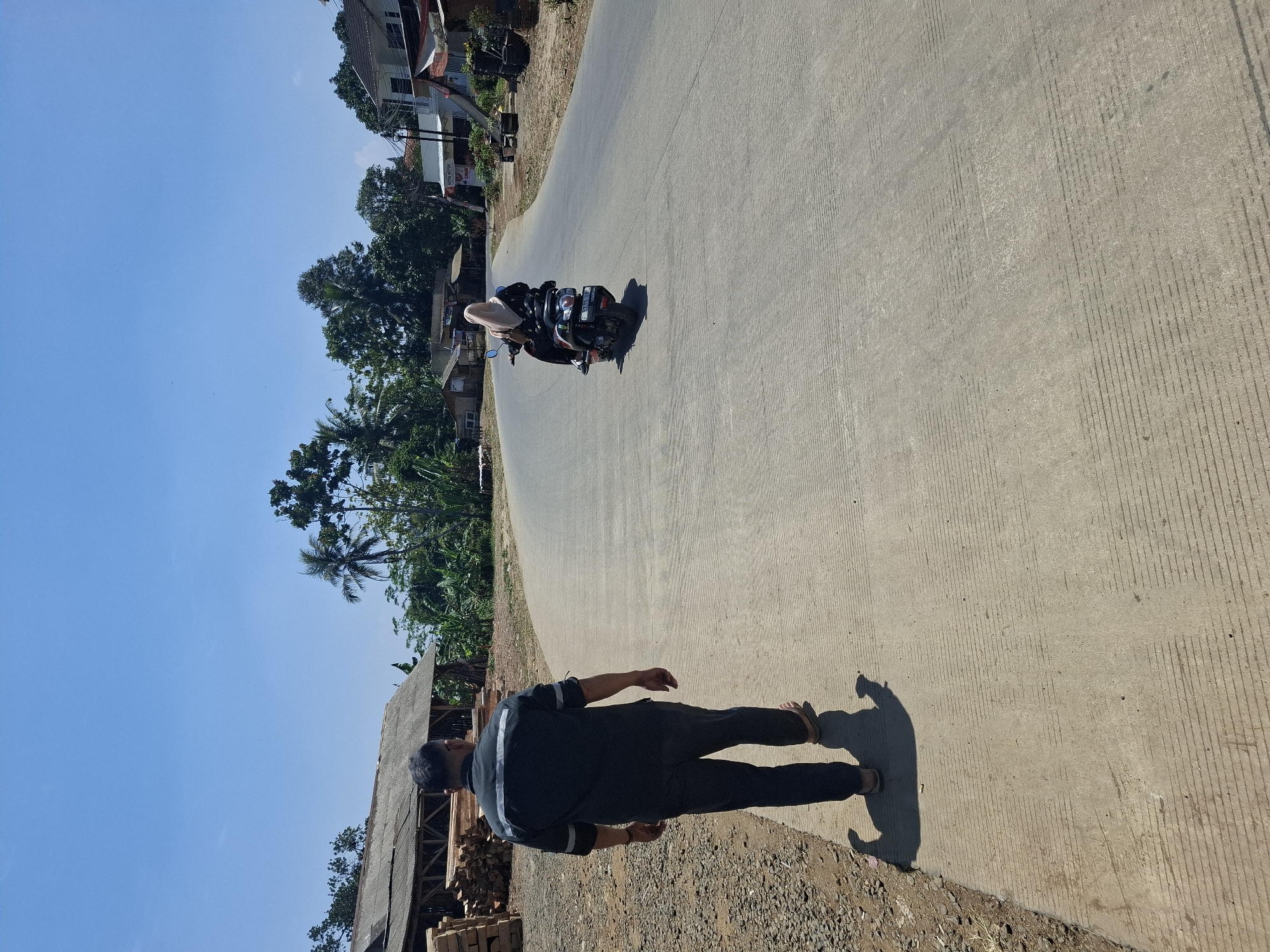 In the city of Ciheulang Tonggoh, Indonesia, on a clear day with a bright blue sky, a man walks away from the camera on a paved road. He is wearing a dark long-sleeved shirt with reflective stripes on the shoulders and arms, and dark pants. He is also wearing sandals. To his left, a pile of lumber is stacked outside a wooden structure with a corrugated metal roof. Further down the road, a woman wearing a hijab and a patterned dress rides a scooter. The road curves to the right, and on the right side of the road, there are houses and shops, including one with a sign that reads "ASTRA MOTOR". Lush green trees and vegetation are visible throughout the scene, with some palm trees. The pavement has textured lines running across it, and shadows of people and objects are cast on the ground.