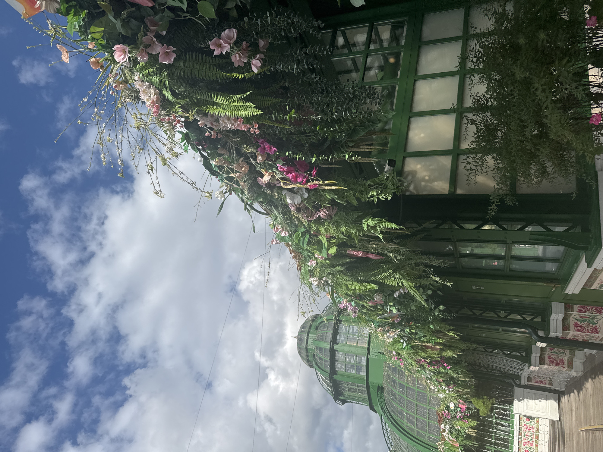 This is a medium shot, taken from a slightly low angle, of a green building adorned with abundant floral and foliage decorations. The building features a large, domed structure in the background, also green, with glass panels. The foreground and right side of the image show a section of the building with green window frames and frosted glass panes. A dense arrangement of artificial flowers and greenery cascades over the roofline and down the side of the building, featuring various shades of pink, white, and green. The sky above is a bright blue with scattered white clouds, indicating daytime. In the distance, a wooden walkway is visible, and the lower part of the building's facade has decorative tiling. There are no people or animals visible in the image.