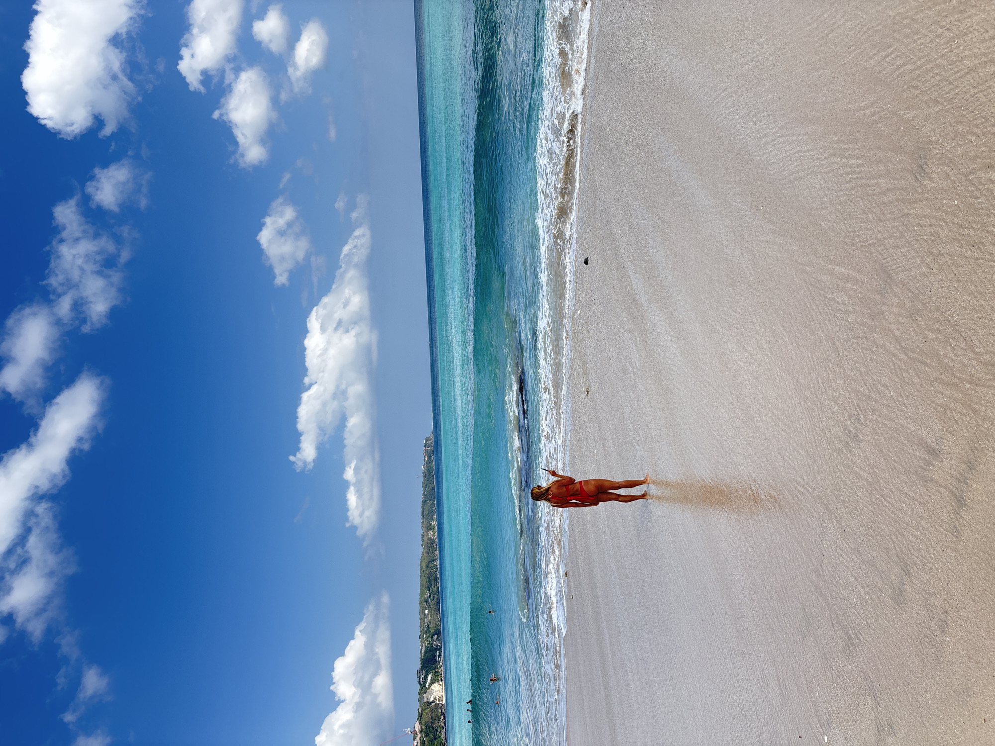 A woman in an orange swimsuit stands on a sandy beach, facing away from the camera and towards the ocean. She is holding a phone in her right hand. The beach is wet, with ripples in the sand indicating receding waves. The ocean water is a vibrant turquoise, with breaking waves near the shore and calm water further out. In the distance, cliffs with vegetation and some buildings are visible along the coastline. The sky is bright blue with scattered white clouds. Several people are in the water in the distance, some appearing to be swimming. The scene suggests a sunny day at a tropical beach location, likely in Kuta Selatan, Indonesia, known for its picturesque coastlines and surf spots.